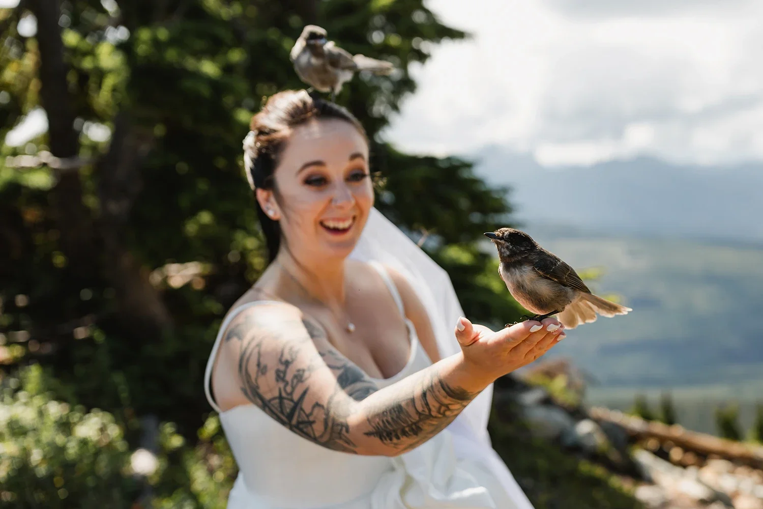 A woman in a white dress holding a bird on her outstretched hand outdoors, smiling with a blurred background of trees and mountains.