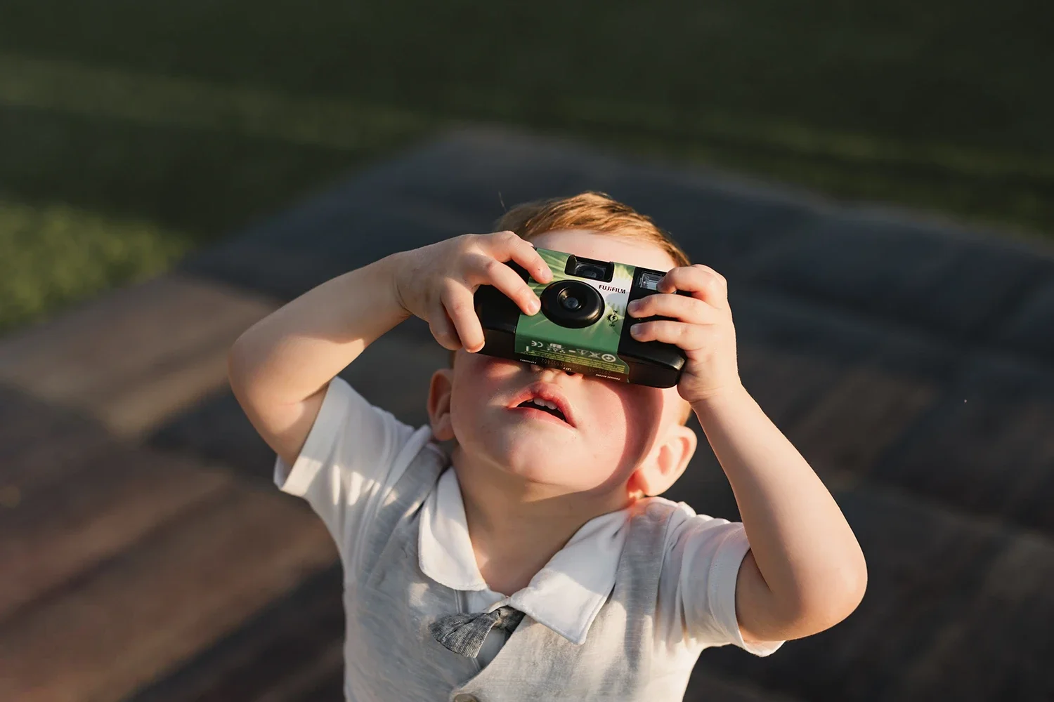 A young boy with light skin and red hair holding a camera up to his face, looking through the viewfinder, outdoors on a sunny day.