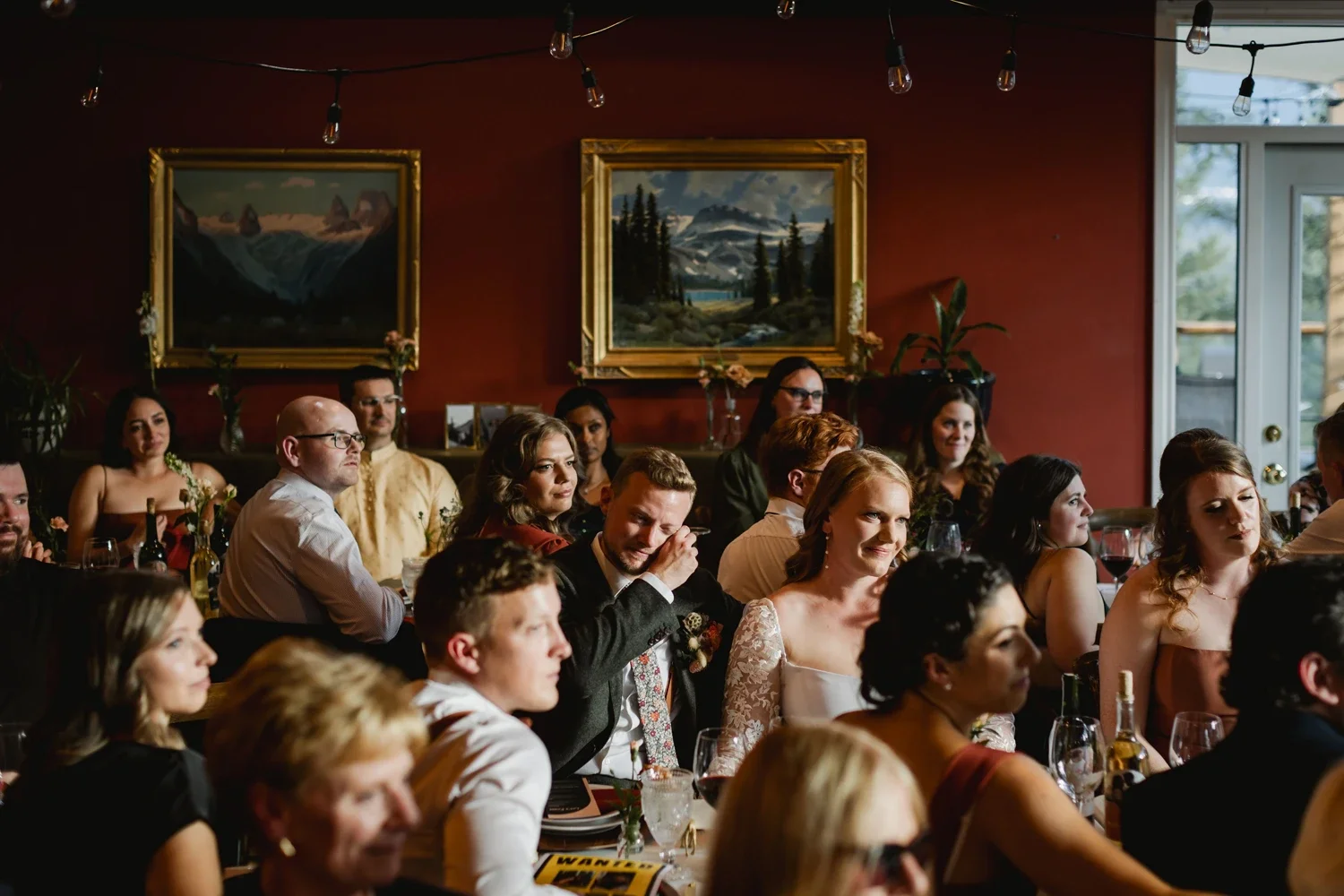 People gathered at a wedding reception, seated at tables in a warmly lit room with red walls, framed landscape paintings, and string lights overhead.