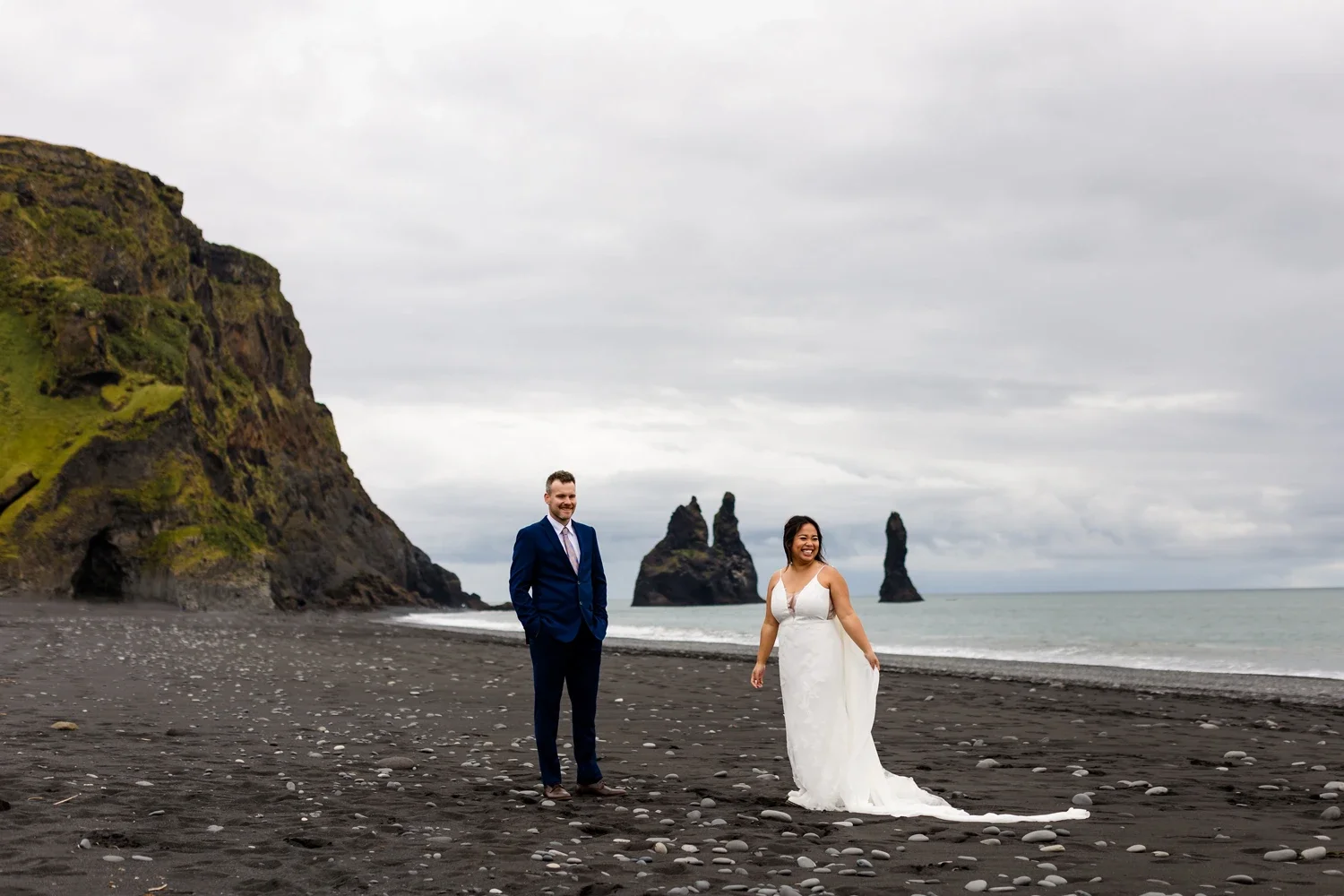 A newlywed couple in wedding attire stands on a black sand beach with rocky cliffs and sea stacks in the background, under a cloudy sky.