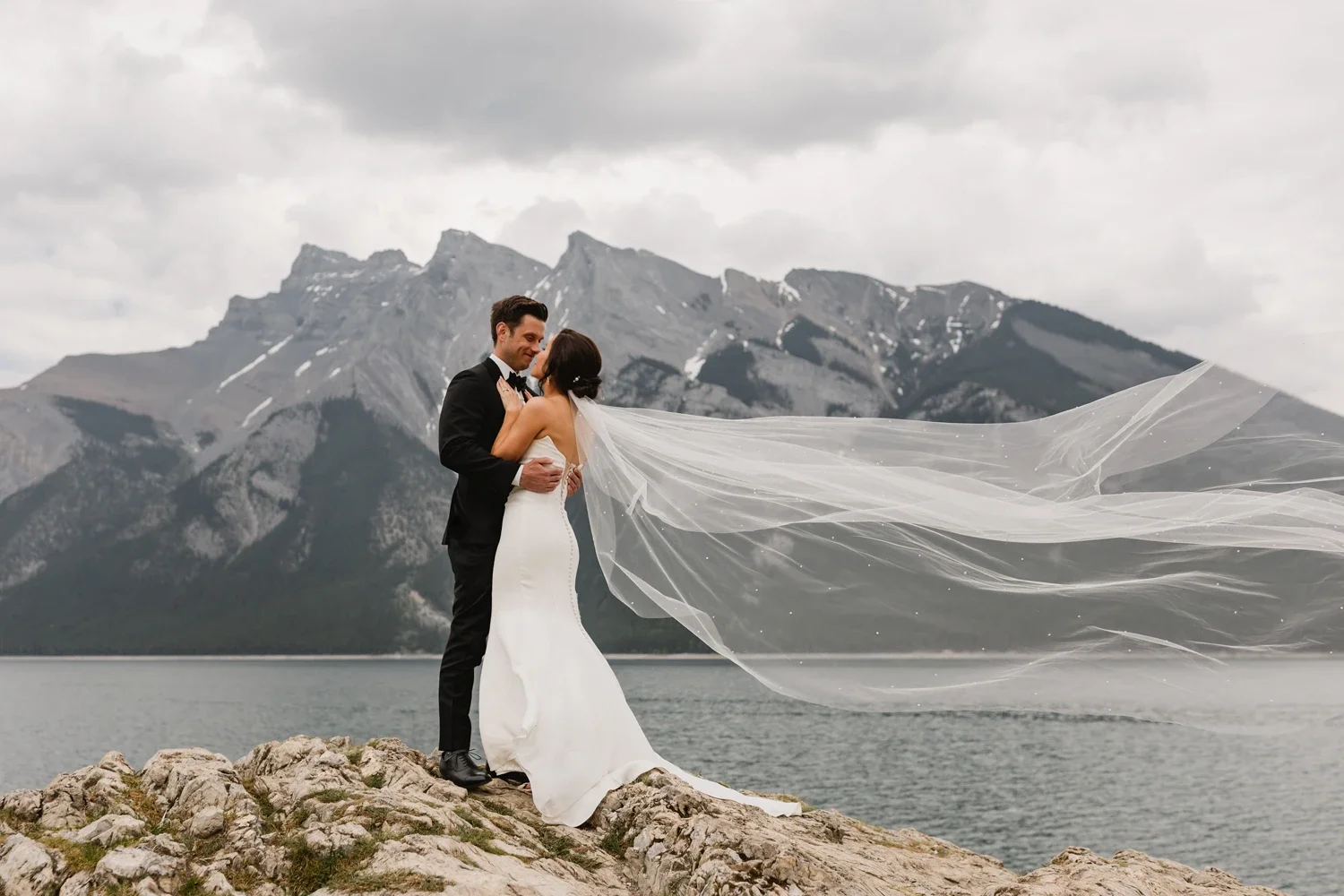 A bride and groom sharing a dance on a rocky shore with mountains and a cloudy sky in the background.