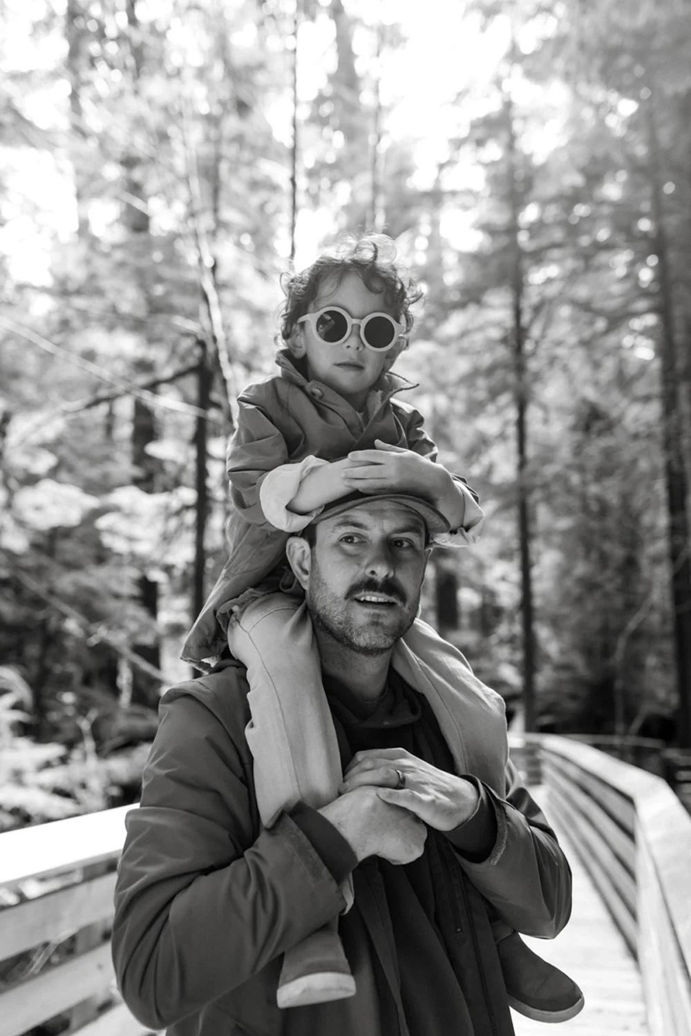 A man carrying a child on his shoulders walking on a forest trail in black and white.