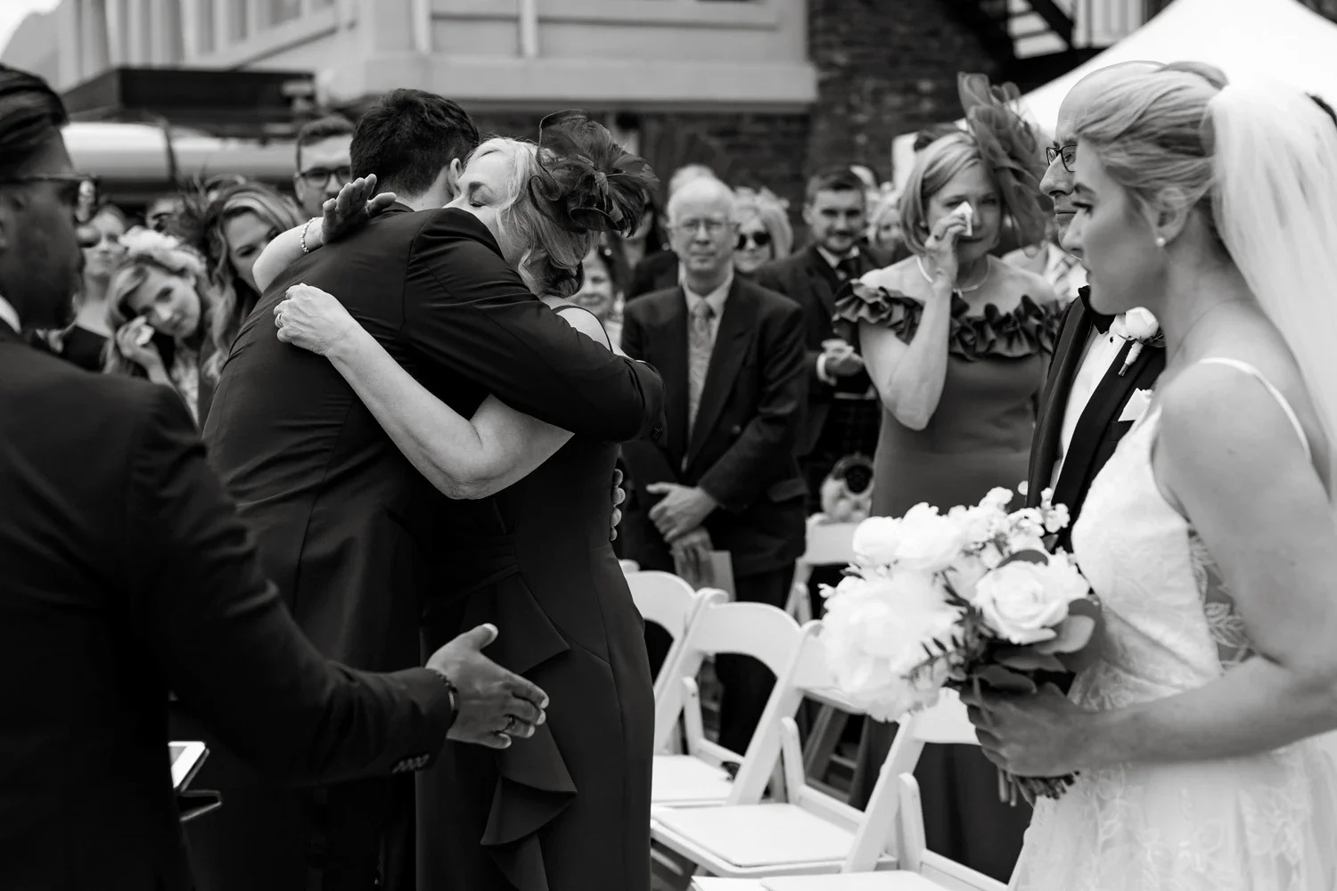 A black and white photo of a wedding ceremony with a woman in a bridal dress holding a bouquet, and several guests, including a man hugging a woman, with more guests in the background.