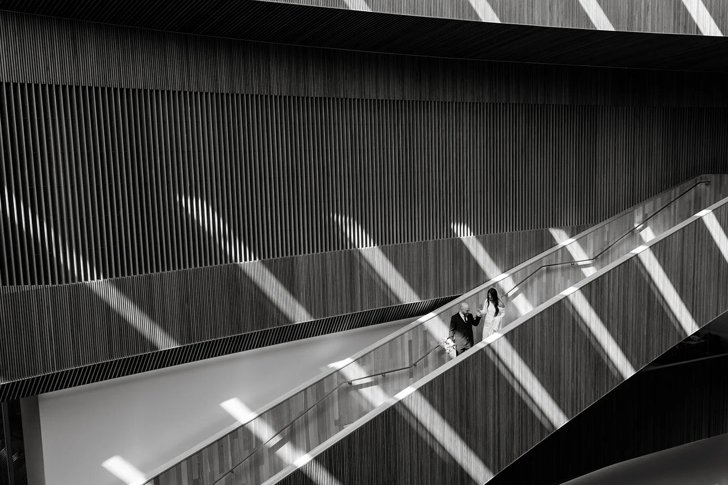 A black-and-white photo of a modern, multi-level building interior with a sweeping staircase. Two people are walking up the stairs, one of whom appears to be a bride holding a bouquet, accompanied by a man in formal attire. The interior features vertical wooden slats on the walls and dynamic patterning of light and shadows cast by windows.