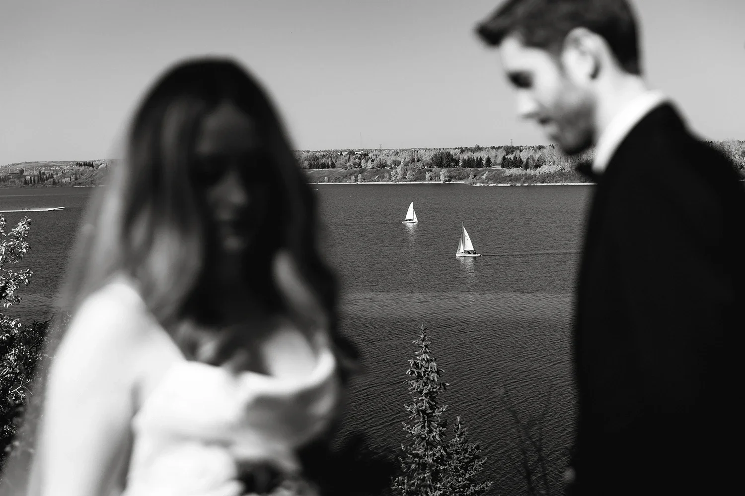 Blurry black and white photo of a woman and man in formal attire near a body of water with sailboats and a distant shoreline.