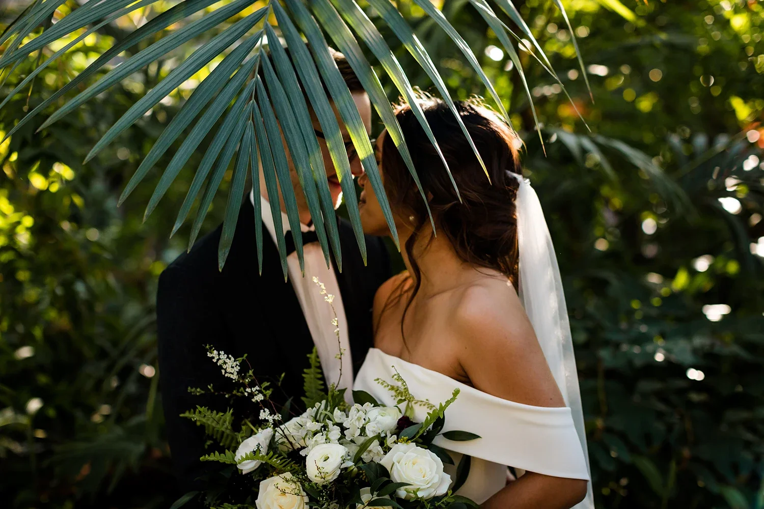 A bride and groom sharing an intimate moment in a lush outdoor setting, partially obscured by tropical leaves, with the bride holding a bouquet of white roses and greenery.
