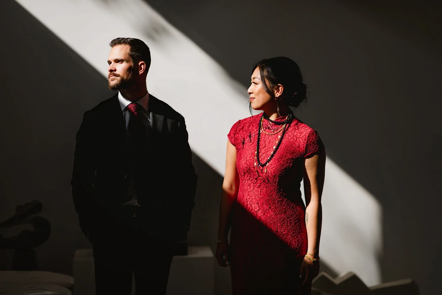 A man in a black suit with a white shirt and red tie, and a woman in a red lace dress with layered necklaces, standing side by side in a room with dramatic lighting and shadows.