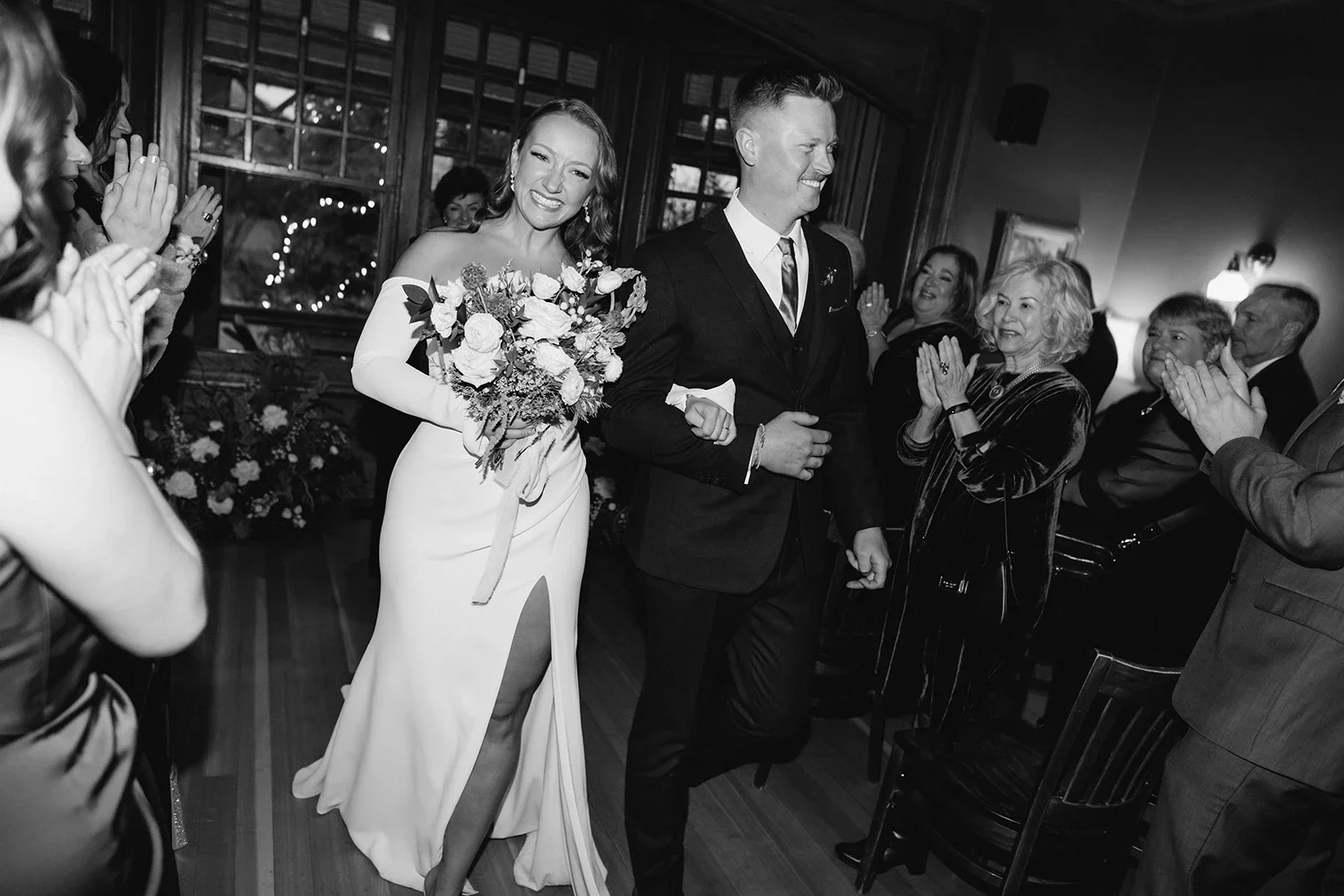 Black and white photo of a wedding reception showing a bride and groom walking arm in arm through a crowd of guests who are clapping and smiling. The bride is holding a bouquet of flowers and wearing a white dress with a slit. The groom is in a dark suit and tie. The atmosphere appears joyful and celebratory.