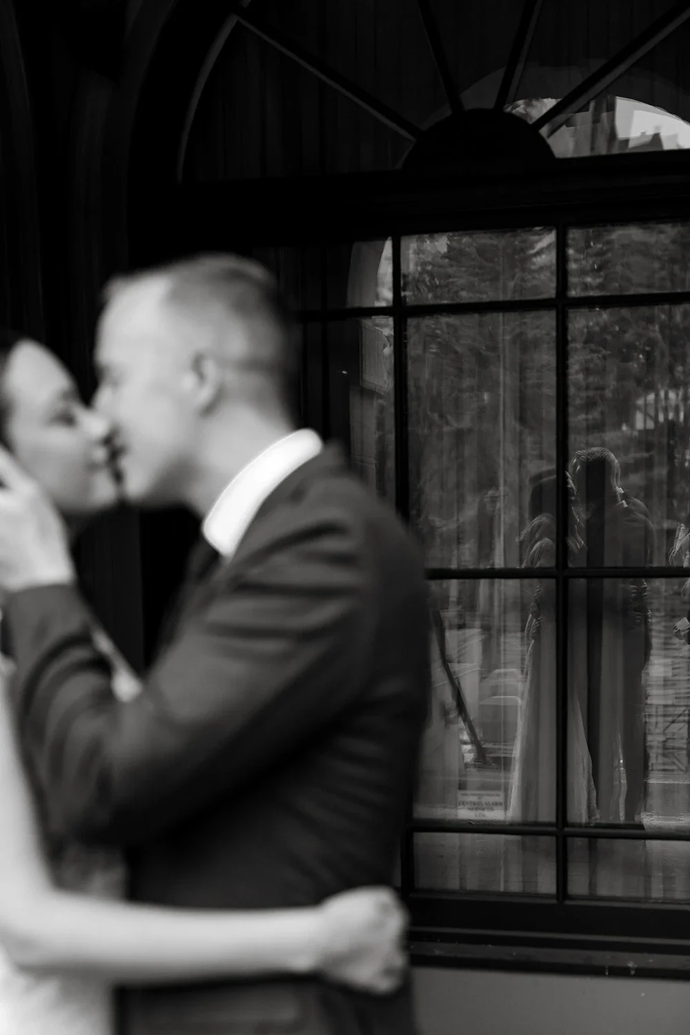 A couple on their wedding day sharing a kiss indoors, with a large window behind them reflecting people outside.