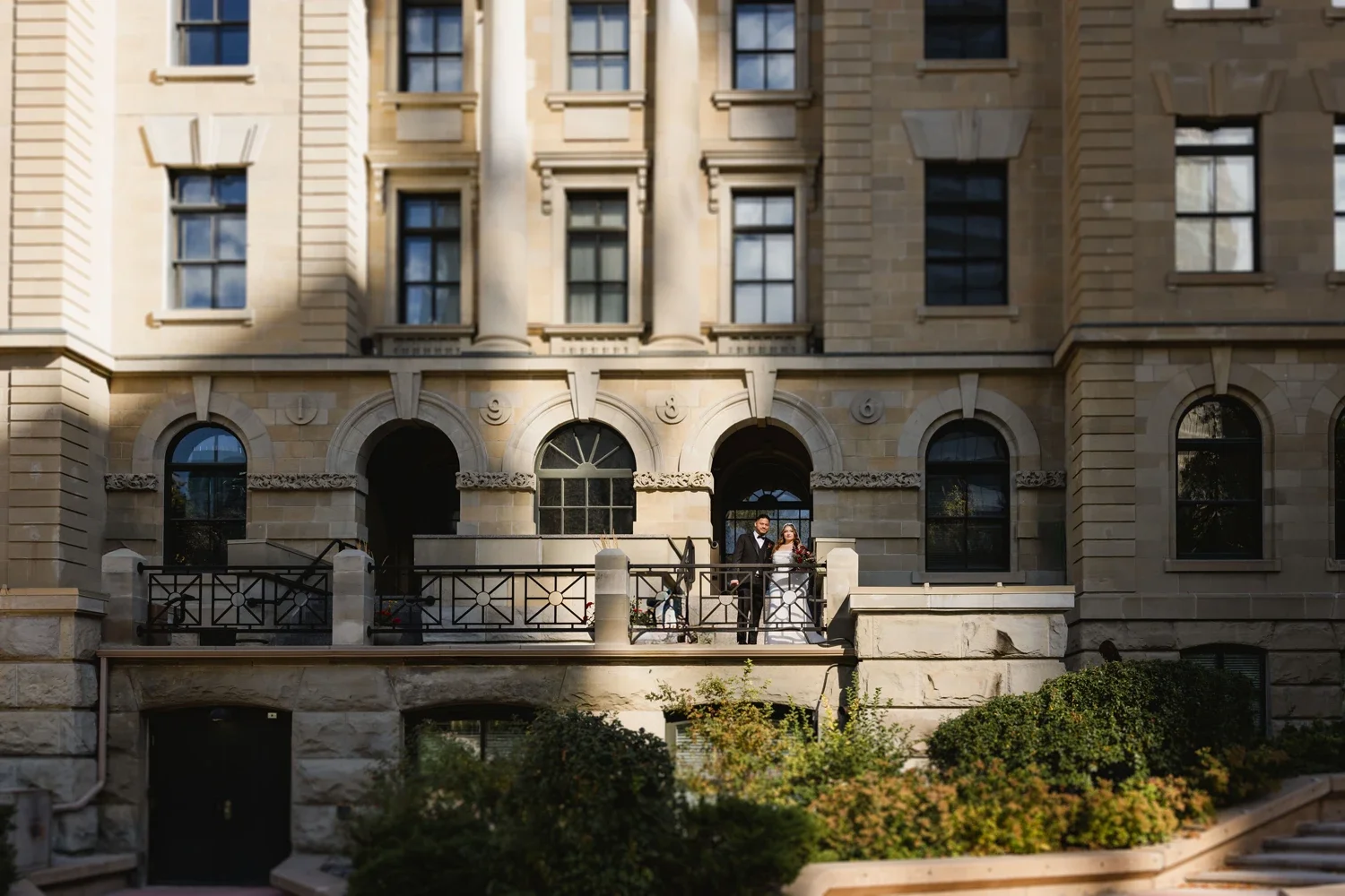 A couple dressed in wedding attire standing on a balcony of a historic stone building with arched windows and ornate architectural details, greenery in the foreground.