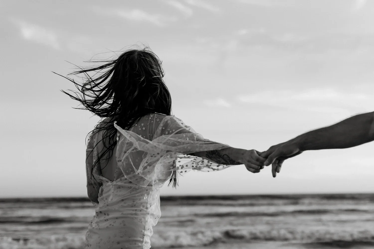 A woman at the beach holding hands with someone, with wind blowing her hair, in black and white.
