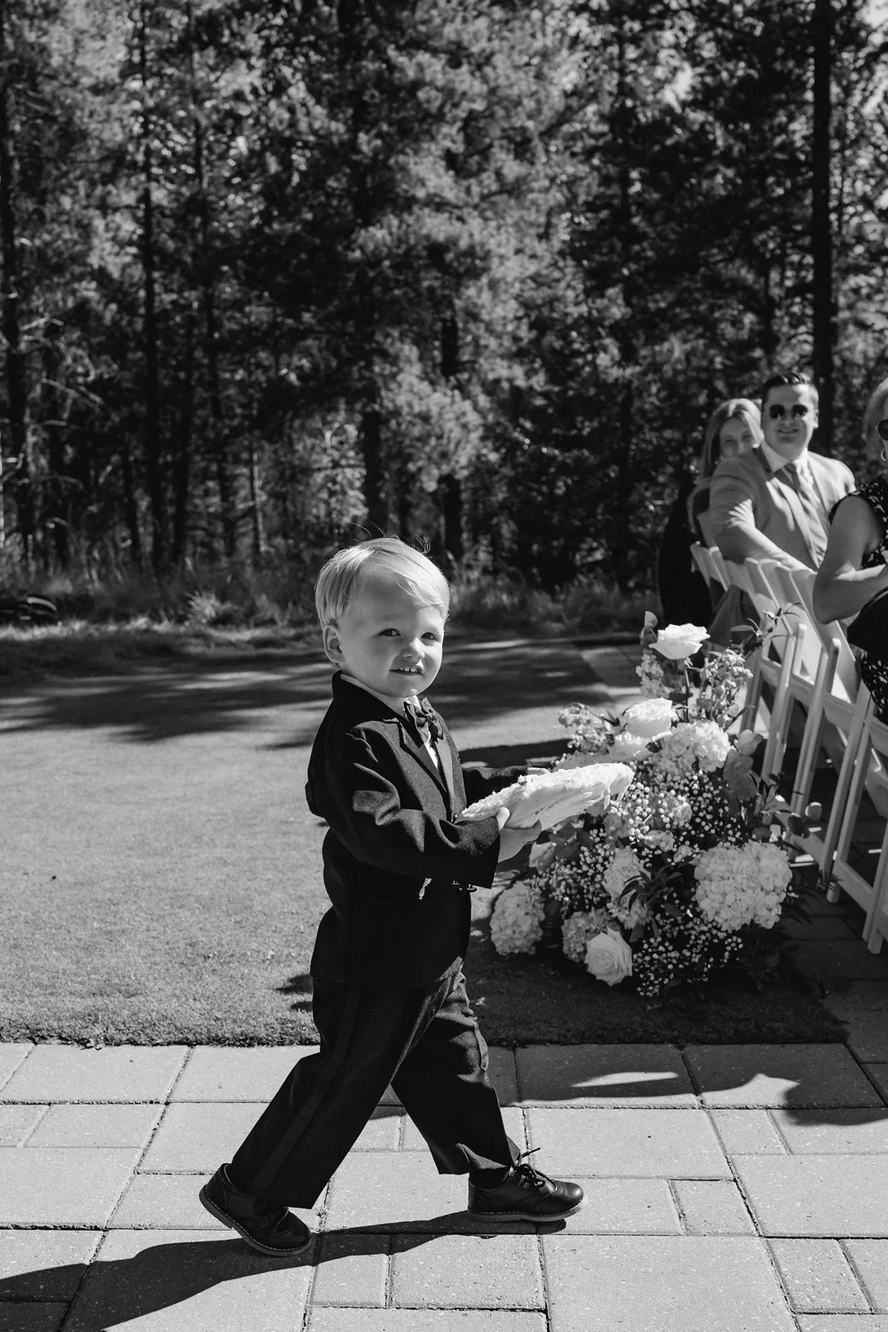 A young boy dressed in a suit walking outdoors on a paved pathway, holding a cake, at a formal event with seated adults, floral arrangements, and trees in the background.