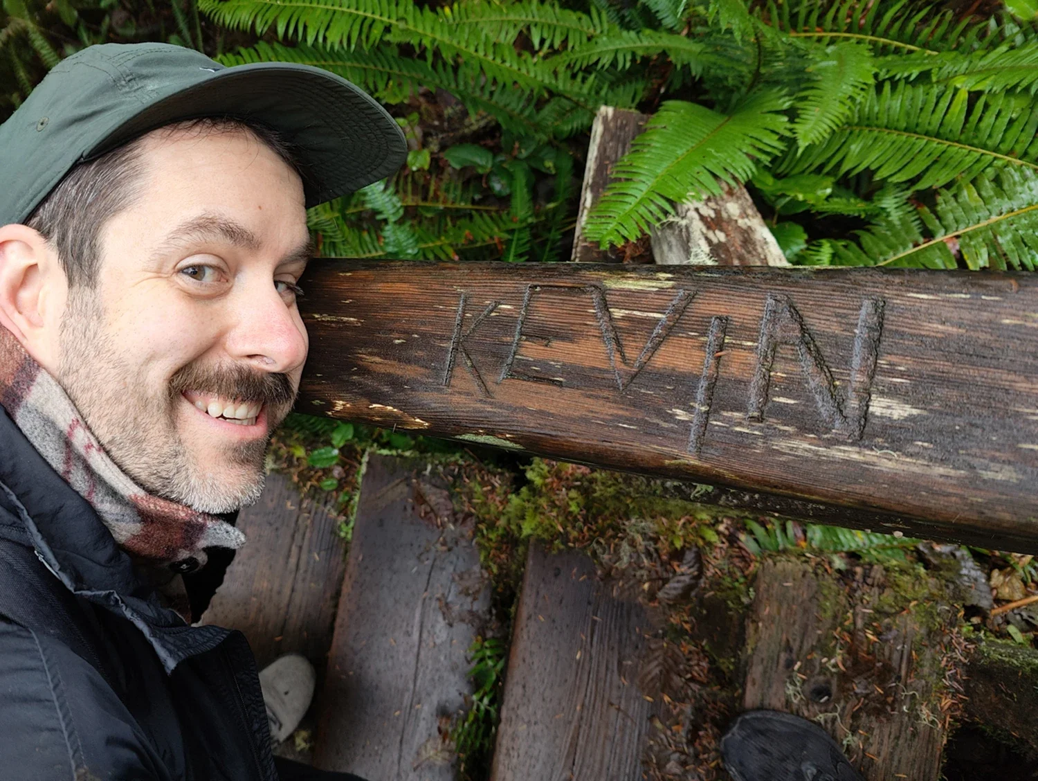 A smiling man wearing a cap and a jacket takes a selfie on a wooded trail during a rainy day. He is standing next to a wooden signpost with the name "KLEVEN" carved into it, surrounded by lush green ferns and foliage.