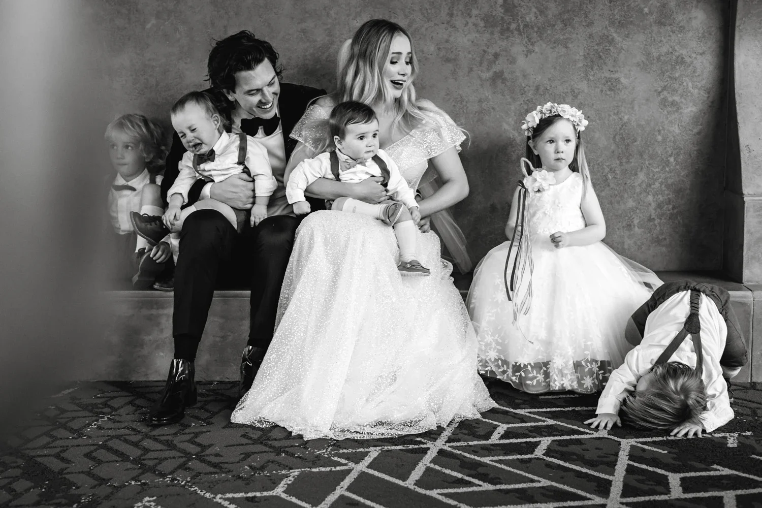 A black-and-white photo of a family at a formal event. A man and woman sit on a bench, holding young children. Four small children are in the scene, with one girl seated to the right wearing a flower crown and a white dress, and another girl is on th