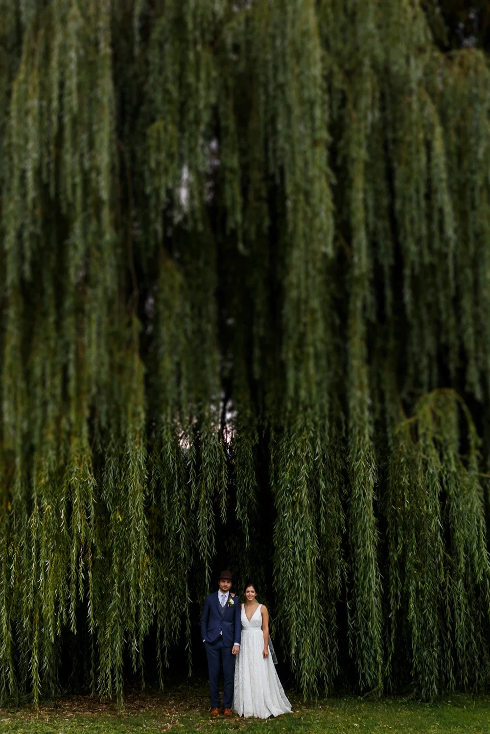 A newlywed couple stands close together outdoors in front of a large weeping willow tree, with the groom wearing a navy suit and the bride in a white wedding dress.