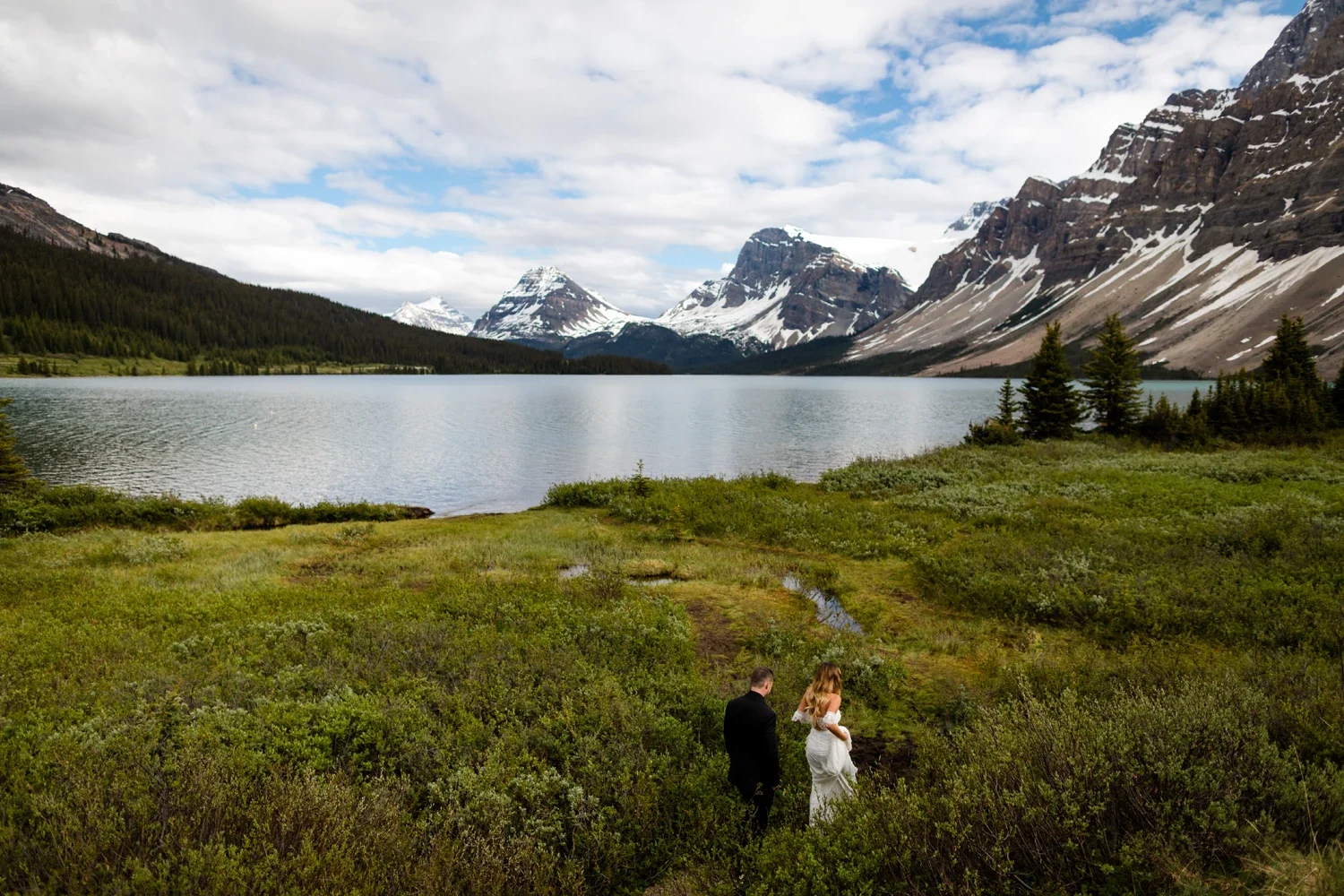 A couple, a man in a black suit and woman in a white dress, walking through green shrubs near a lake with mountains in the background, under a partly cloudy sky.