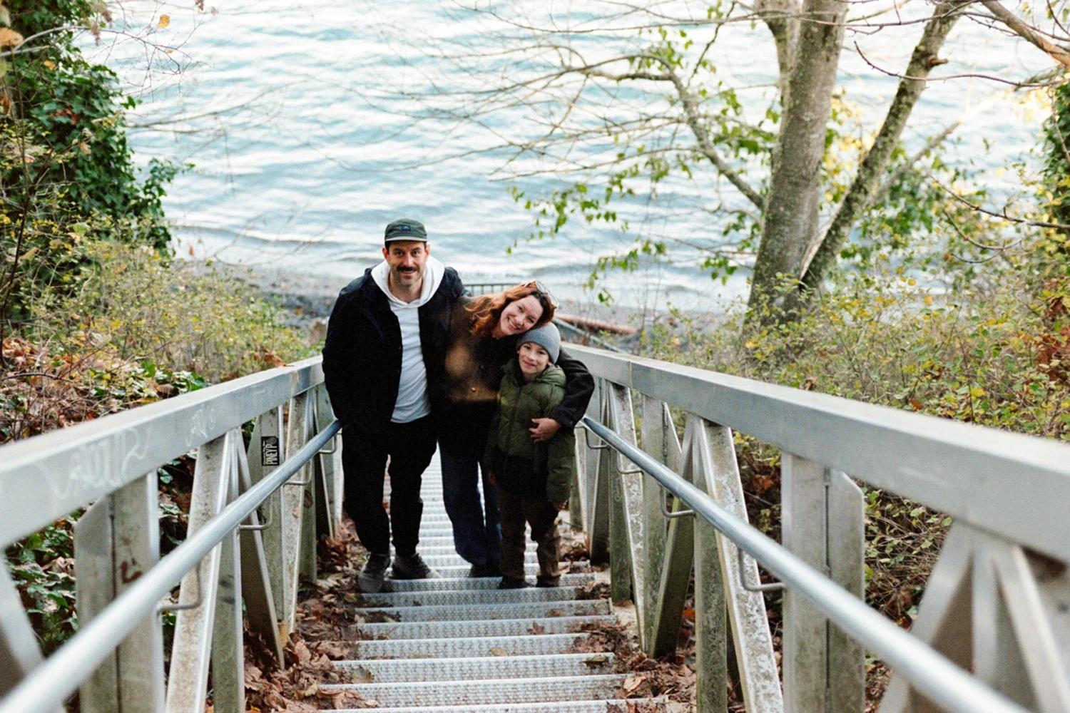 Family of three standing on metal staircase leading down towards a lake, surrounded by trees with autumn leaves.