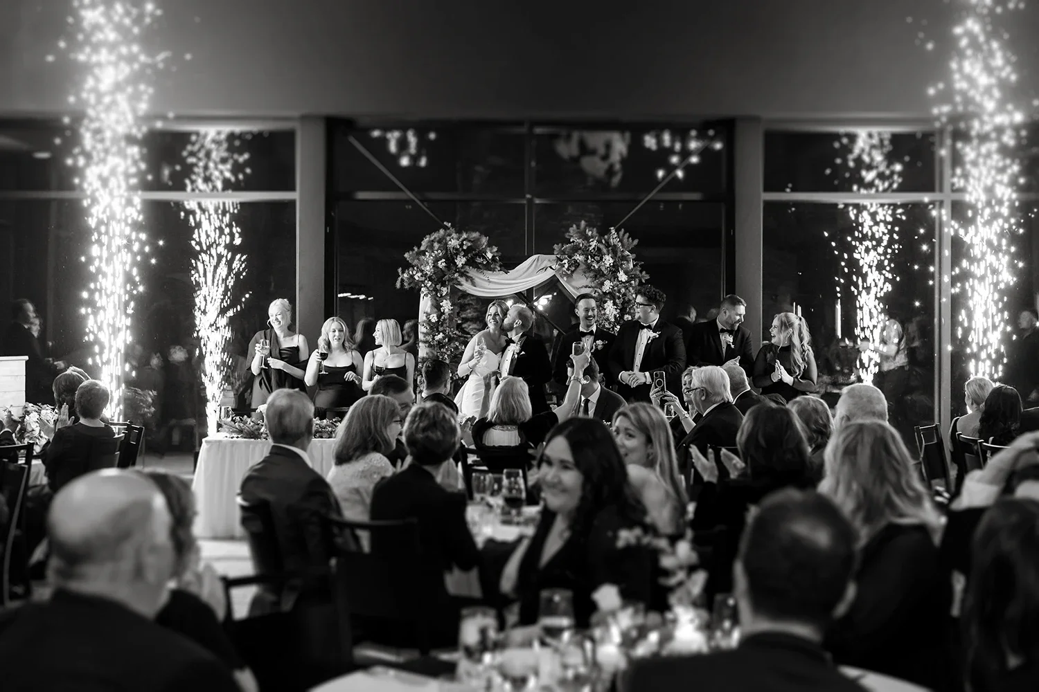 Black and white photo of a wedding reception with a bride and groom kissing on a decorated stage, surrounded by wedding party members and guests, with fireworks and standing guests celebrating.