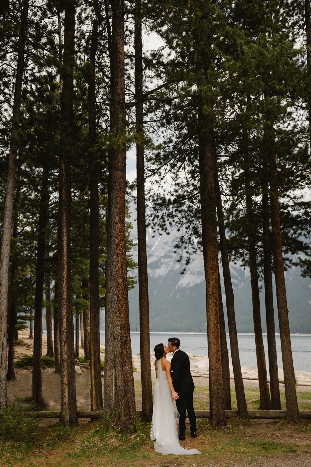 A bride and groom kiss in a forested area near a lake with mountains in the background.