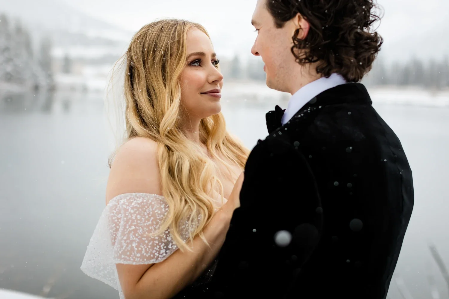 Couple in wedding attire standing close outdoors by a lake in a winter setting, gazing into each other's eyes
