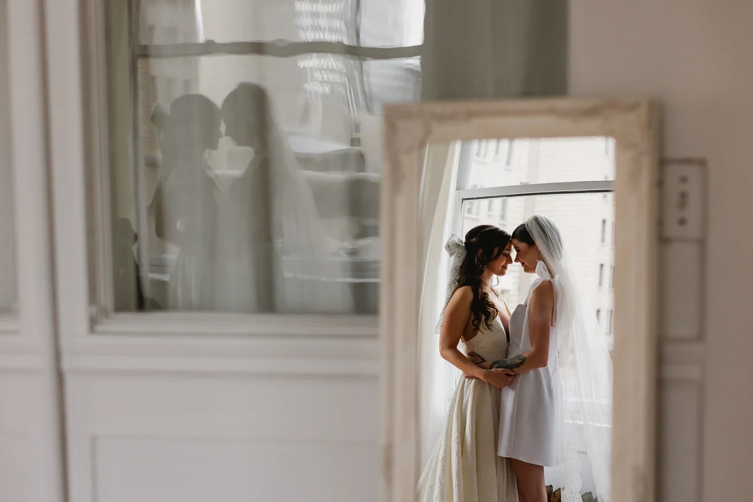 Two women, a bride and her partner, embracing with foreheads touching in front of a window, reflected in a mirror that is partially visible in the foreground.