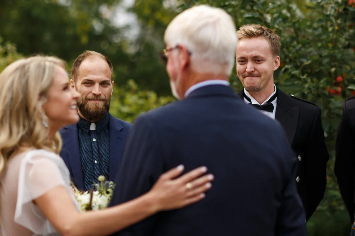 A bride and groom at their wedding ceremony outdoors, surrounded by three men in suits, with one man in black tuxedo, and a background of green trees and red berries.