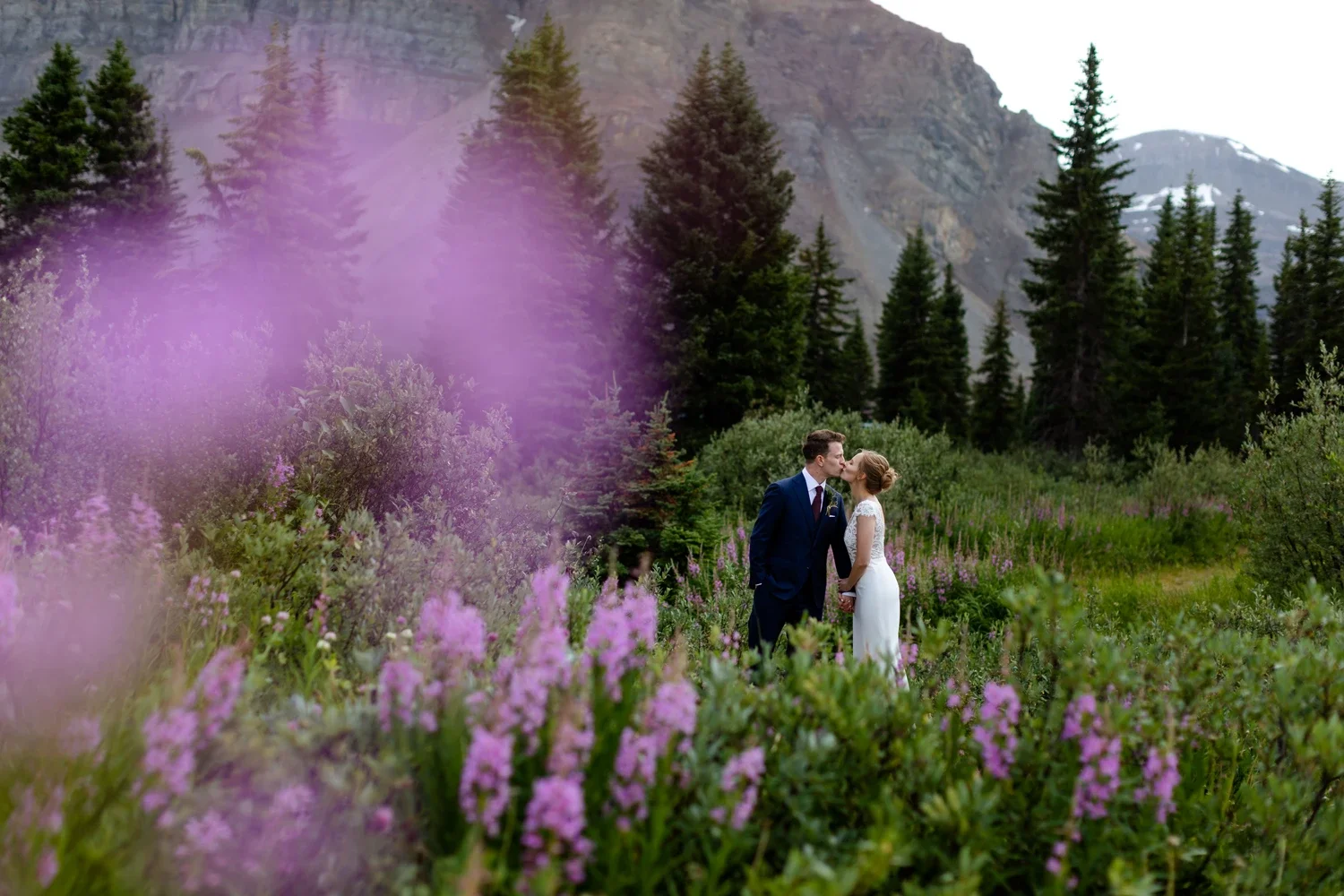 A bride and groom sharing a kiss in a lush, purpleflowered meadow with pine trees and mountain cliffs in the background.