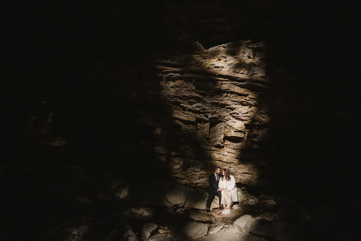 A man and woman sitting close together in a dark cave, illuminated by a beam of light, holding hands and looking at each other.