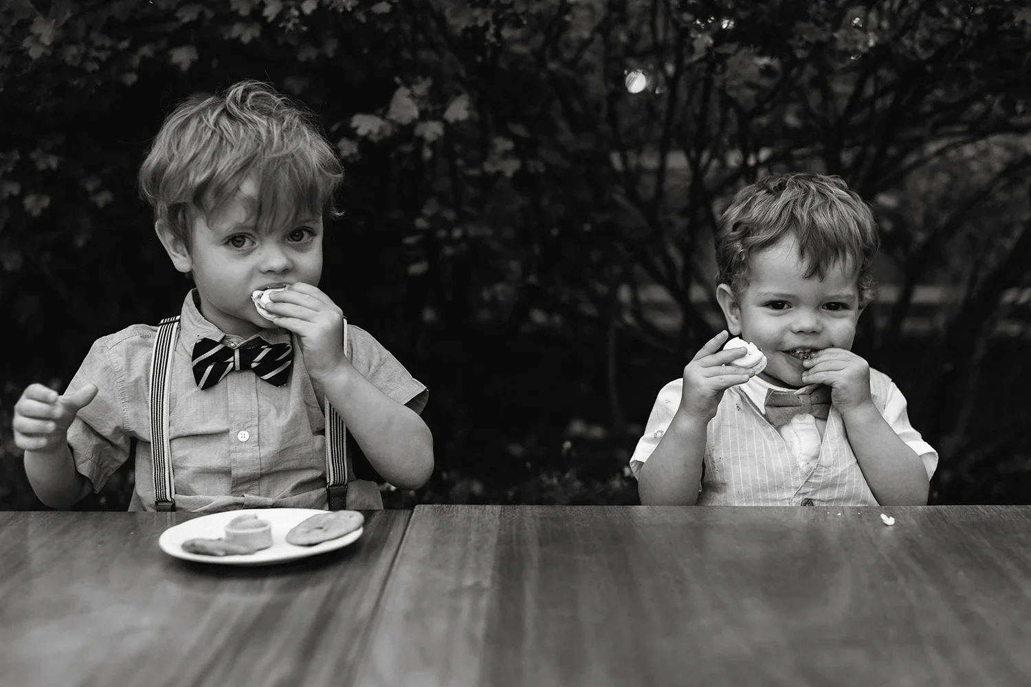 Two young boys sitting at a table, eating cookies, with one wearing a striped bow tie and suspenders, in a black and white photo outdoors.