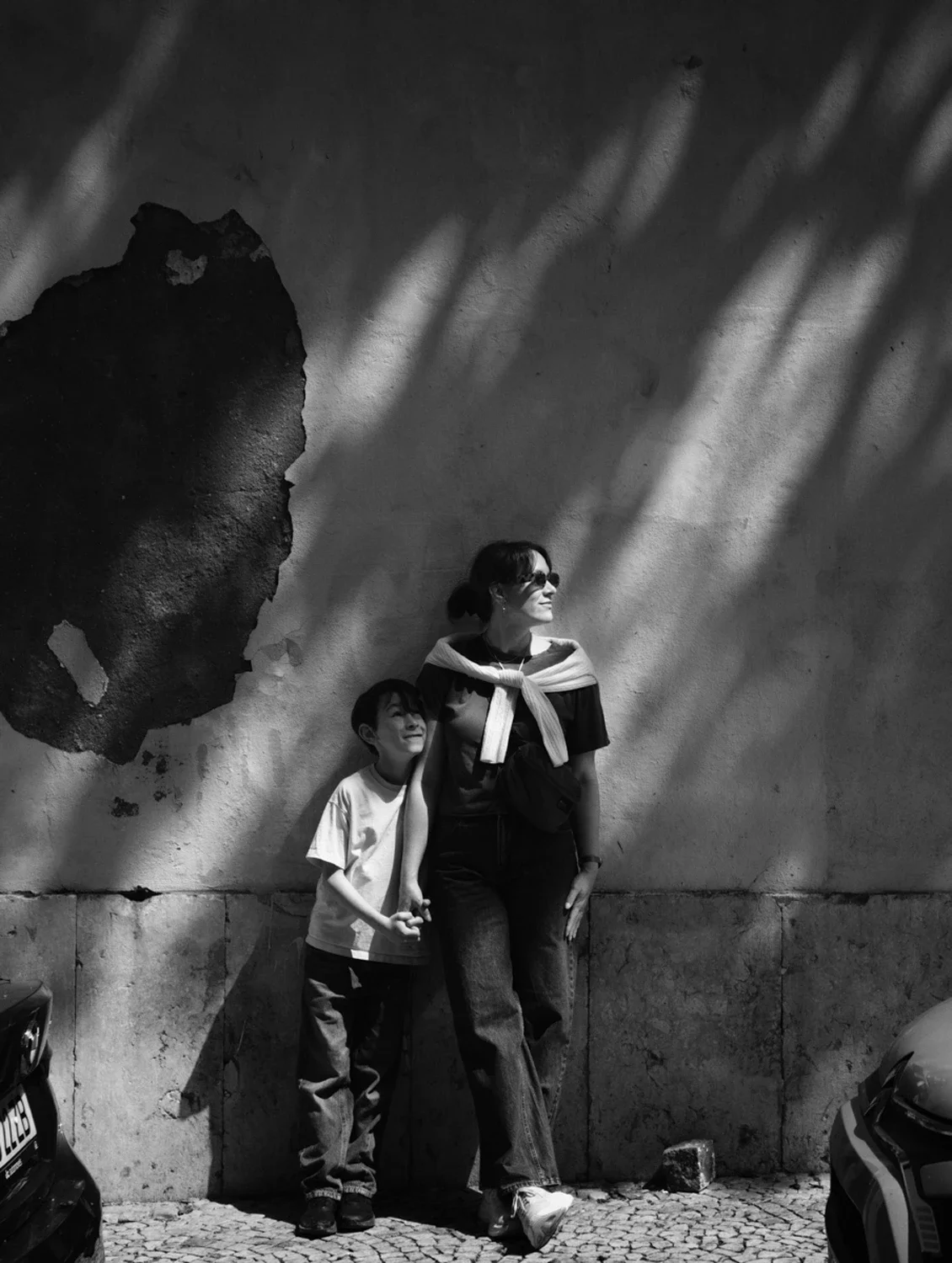 A woman and a young boy standing against a textured wall with sunlight and shadow patterns. The woman is wearing sunglasses, a jacket tied around her shoulders, and casual clothing. The boy is holding her hand and looking up at her, both smiling. The