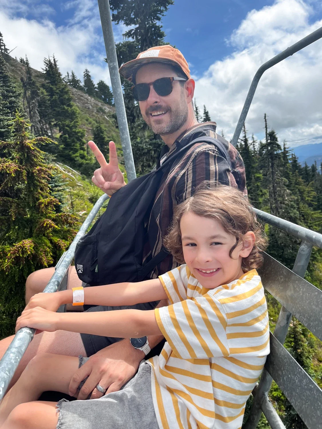 A man and a young girl sitting on a metal mountain coaster track surrounded by pine trees, with mountains and a blue sky in the background. The man is wearing sunglasses, a hat, and a striped shirt, and is making a peace sign. The girl is smiling, we