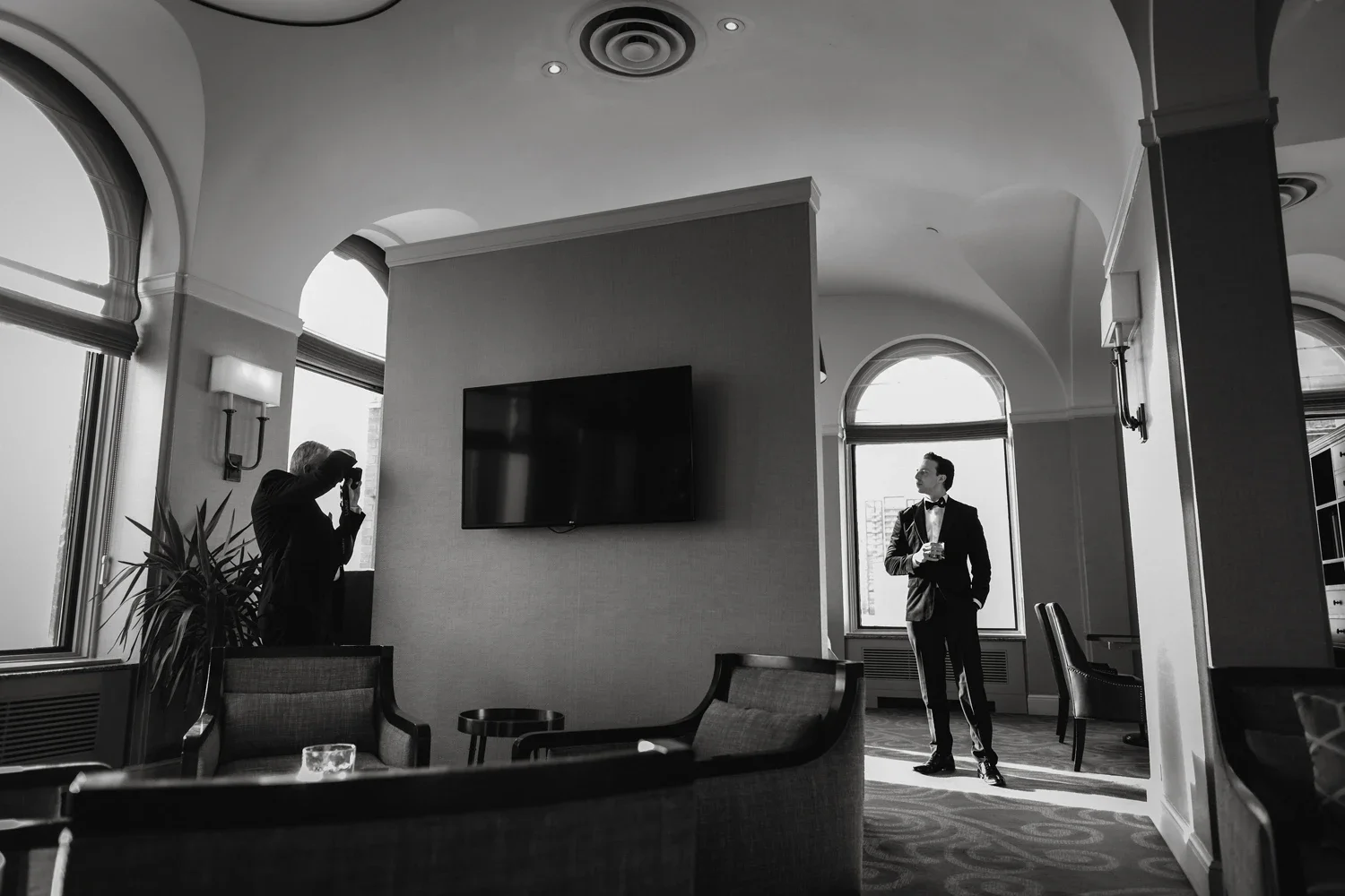 A man in a tuxedo holding a drink is standing near a window, while another person is taking a photograph inside a well-lit, elegant room with arched windows and a large flat-screen TV on the wall.