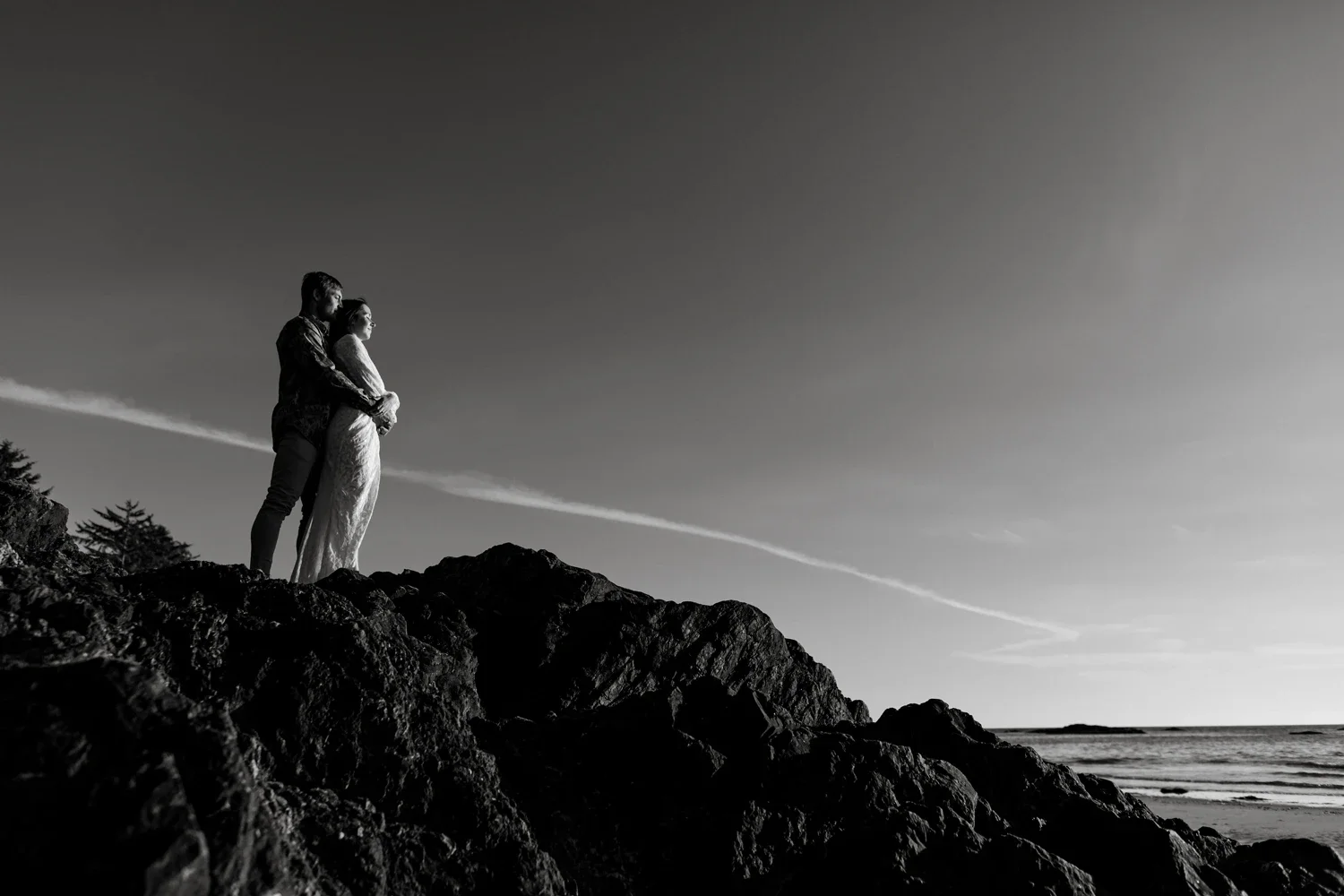 A black-and-white photo of a couple standing on rocky terrain by the ocean, with the man embracing the woman from behind, both looking toward the horizon.