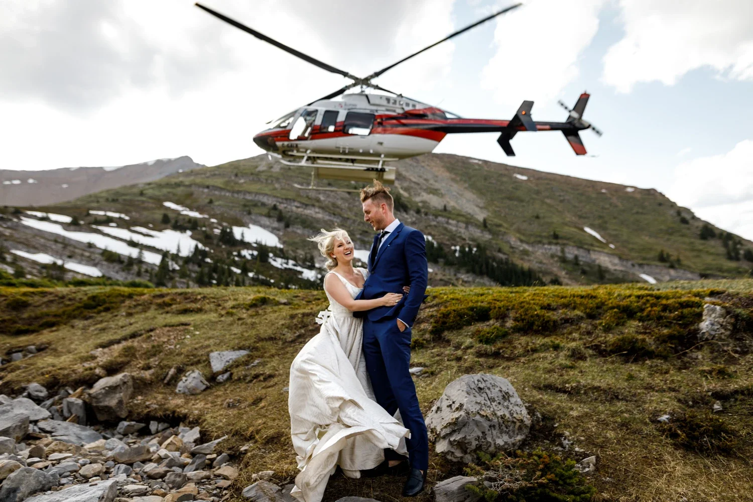 A couple in wedding attire smiling and embracing outdoors in a mountainous area with a helicopter flying overhead.