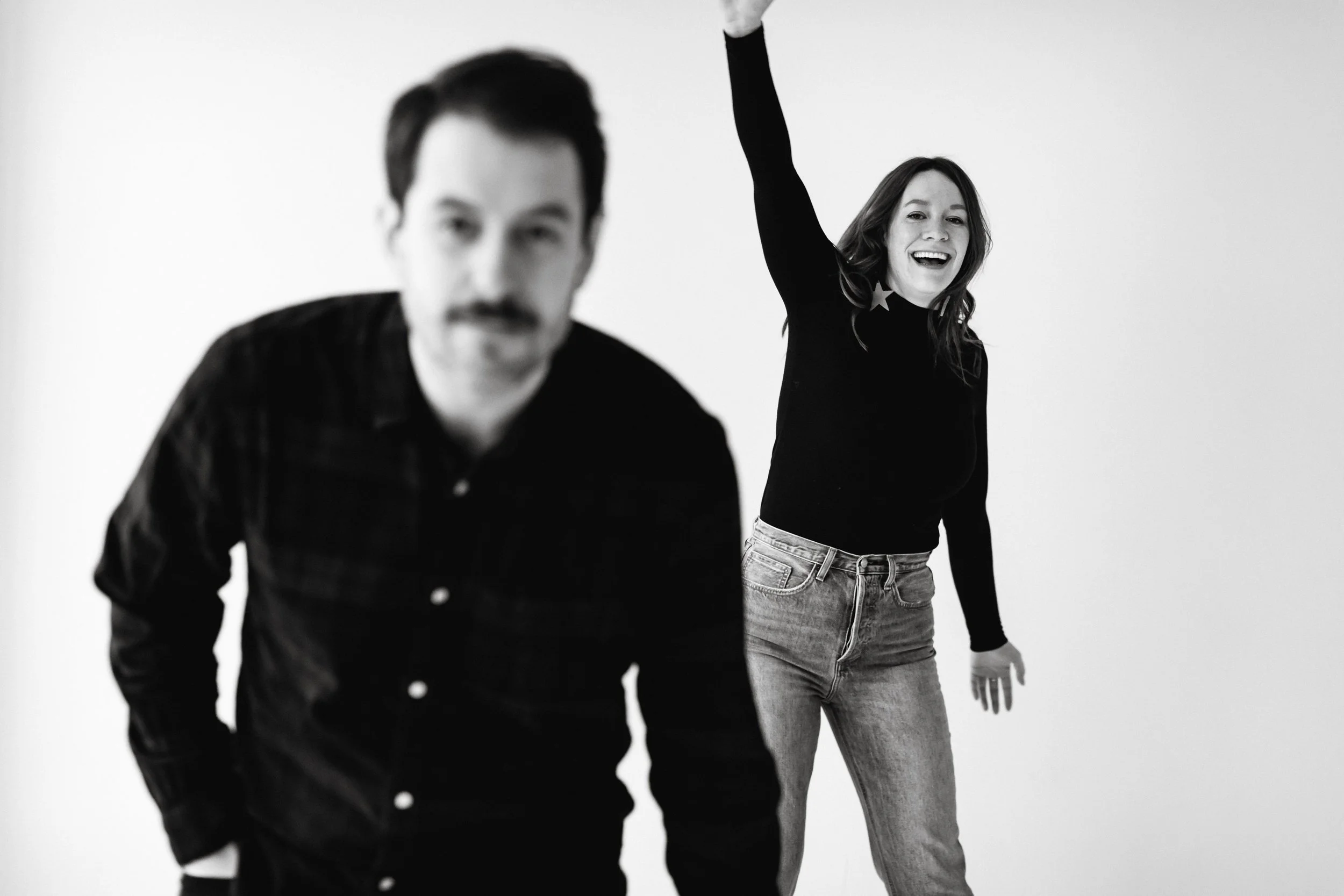 Black and white photo of a man in the foreground out of focus, a woman in the background smiling and raising her right arm with her left hand resting on her hip, wearing a black long sleeve shirt with star details near the collar and jeans.