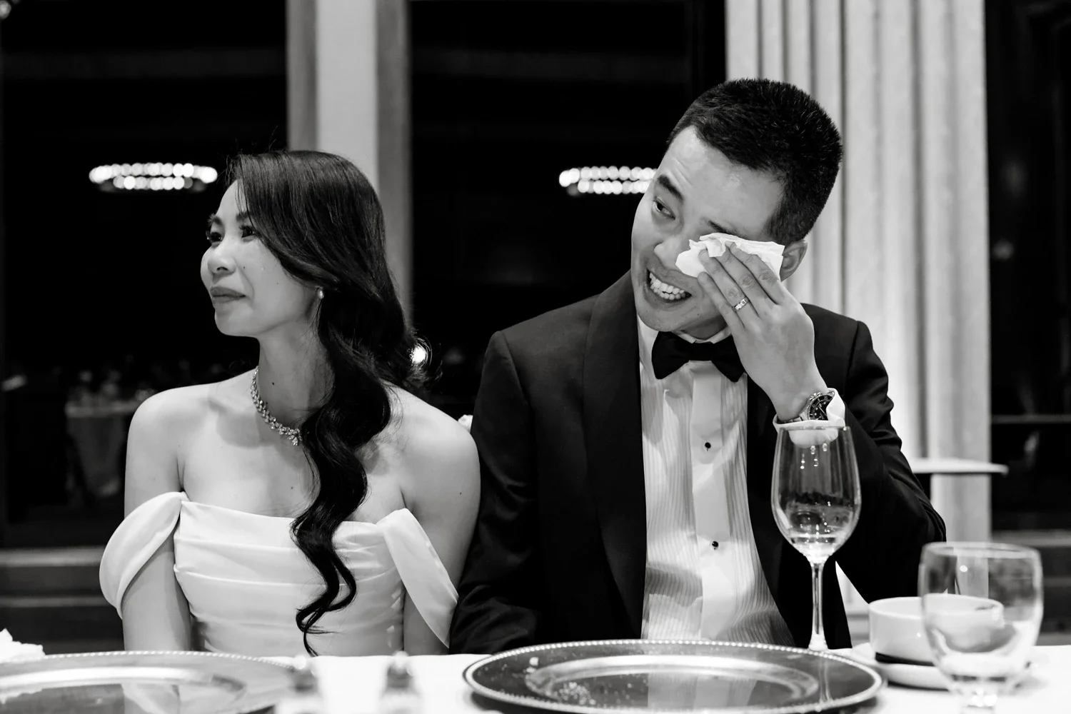 A man in a tuxedo wiping his eye with a tissue while sitting next to a woman in a strapless dress at a formal event.