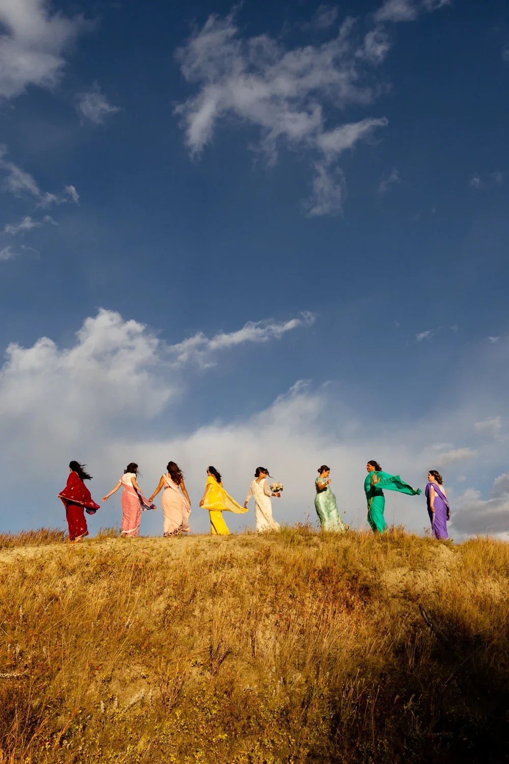 Group of women in colorful dresses walking on a grassy hill under a blue sky with clouds, some holding hands and one holding a bouquet.