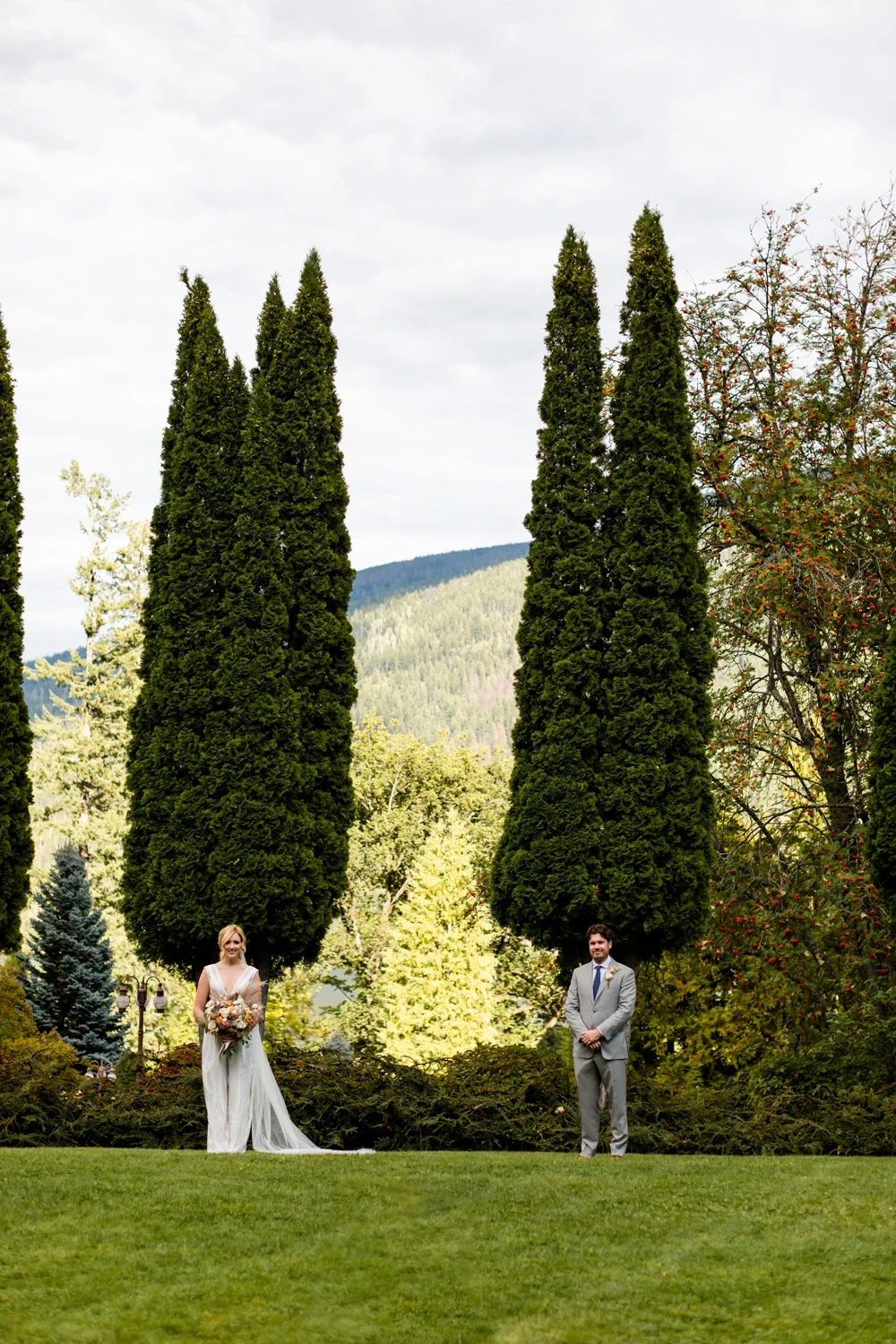 Bride in white wedding dress holding a bouquet standing on the grass, groom in gray suit standing on the grass, tall green trees, and a scenic mountain backdrop under a cloudy sky.