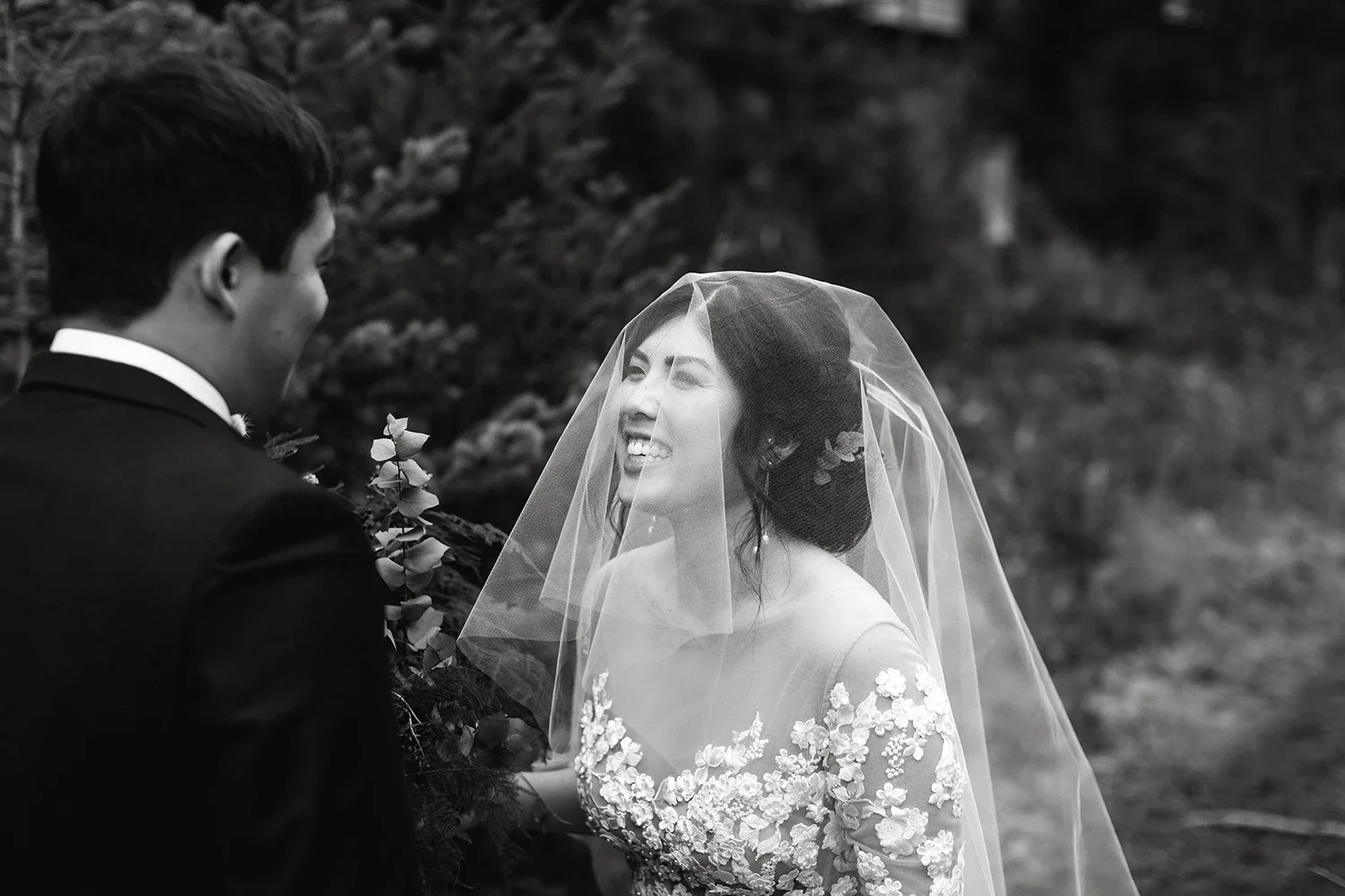A black and white photo of a bride and groom on their wedding day. The bride is smiling, wearing a lace dress and a veil, while the groom is looking at her, dressed in a suit. They are outdoors with a natural background.