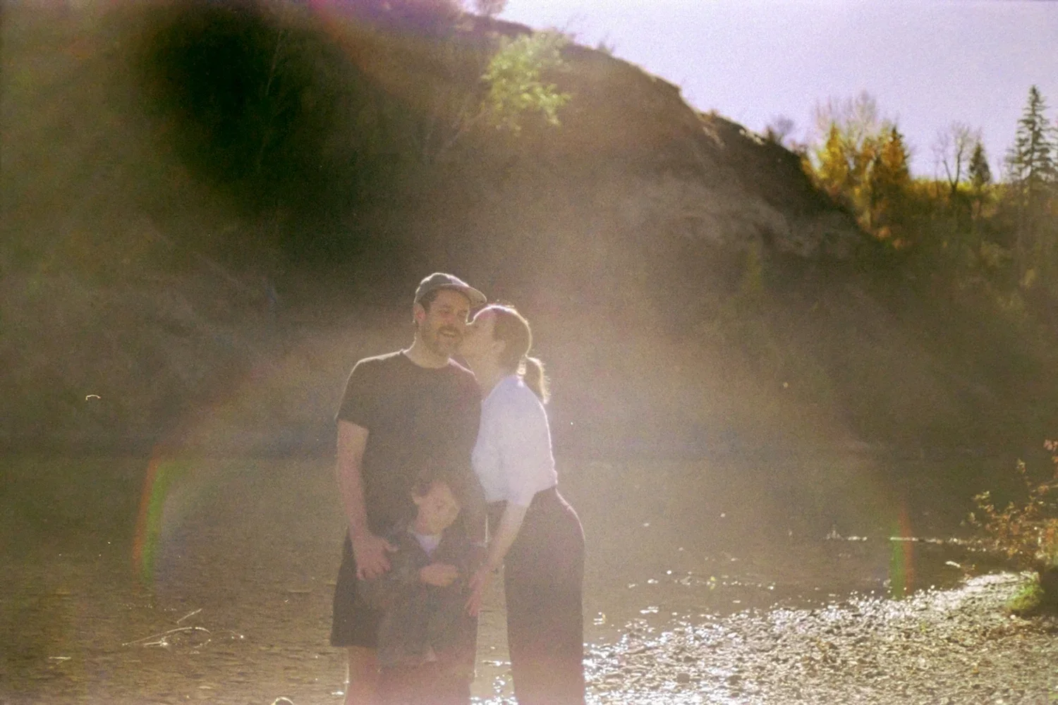 Three people, a man, woman, and child, standing on a sunny riverbank, smiling, with the sun creating lens flares. The background shows hills and trees with autumn foliage.