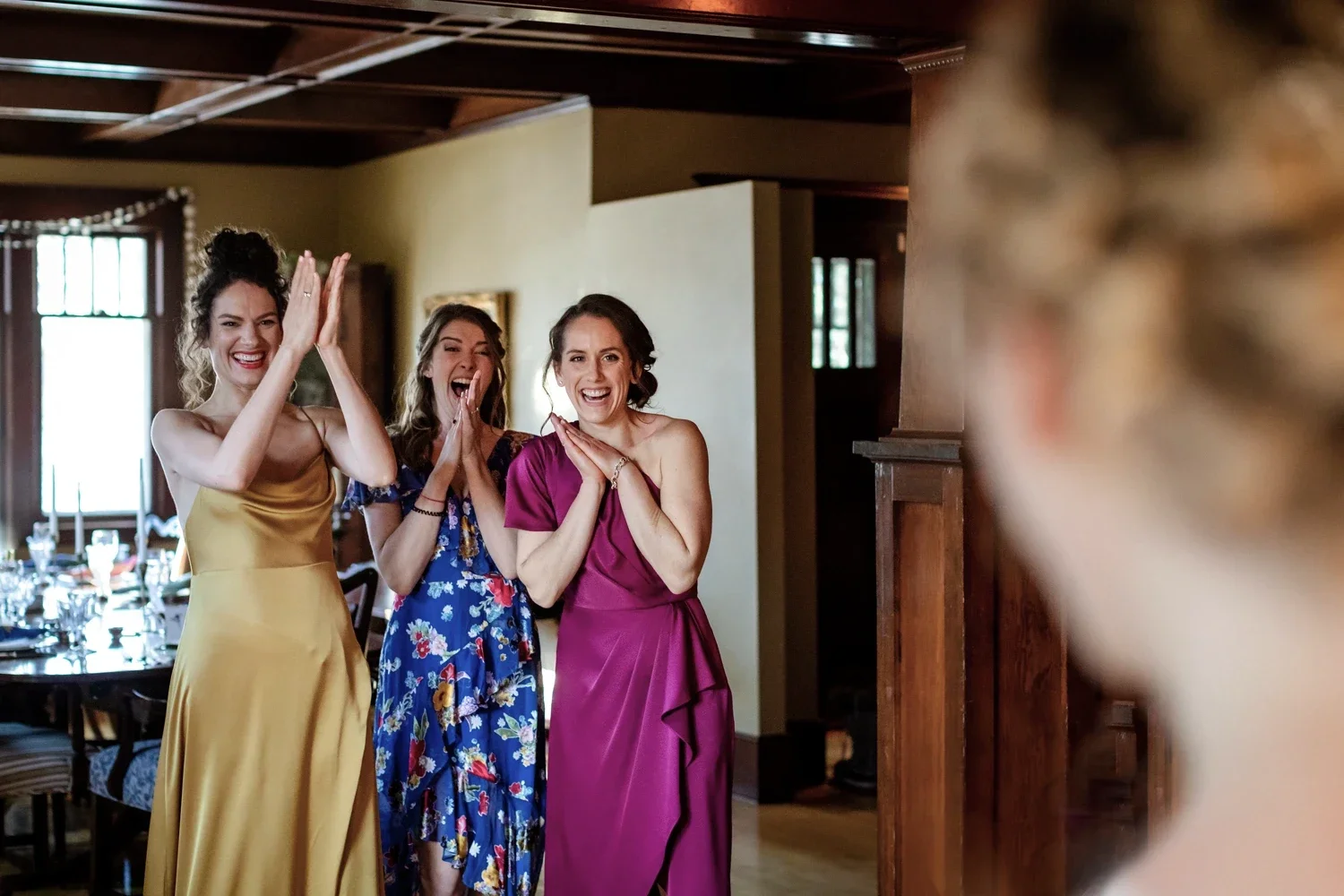 Four women smiling and laughing, wearing colorful dresses, standing indoors in a warmly lit room with tables and chairs in the background.