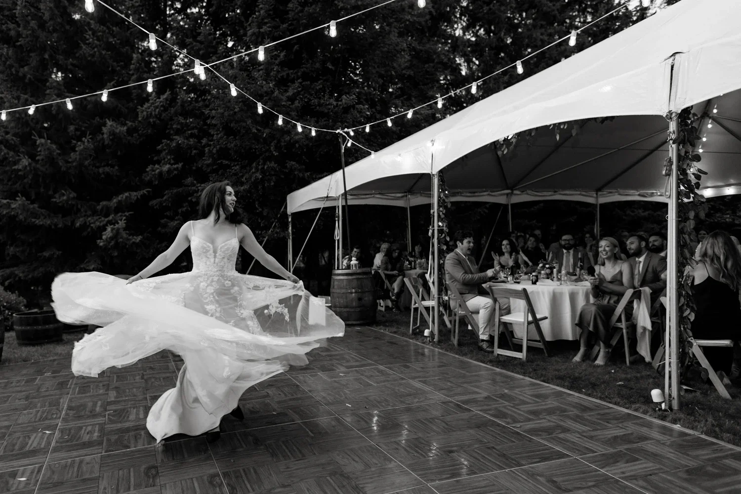A woman in a wedding dress spinning on a dance floor at an outdoor wedding reception, with guests seated at tables under a tent in the background.