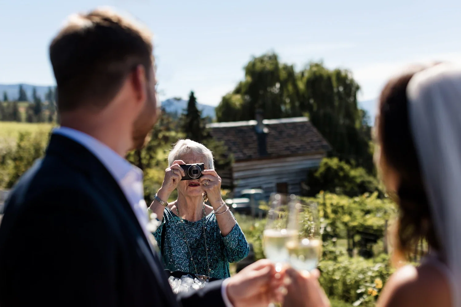A woman with gray hair wearing a patterned blue dress takes a photo with a camera of a couple holding champagne glasses at an outdoor event on a sunny day, with a rustic house and trees in the background.