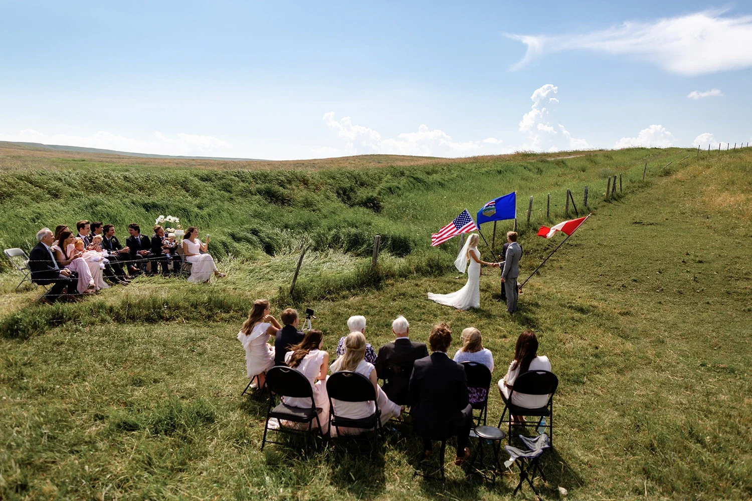 Outdoor wedding ceremony on a grassy hill with a bride and groom exchanging vows, surrounded by guests seated in chairs, with American and state flags in the background.