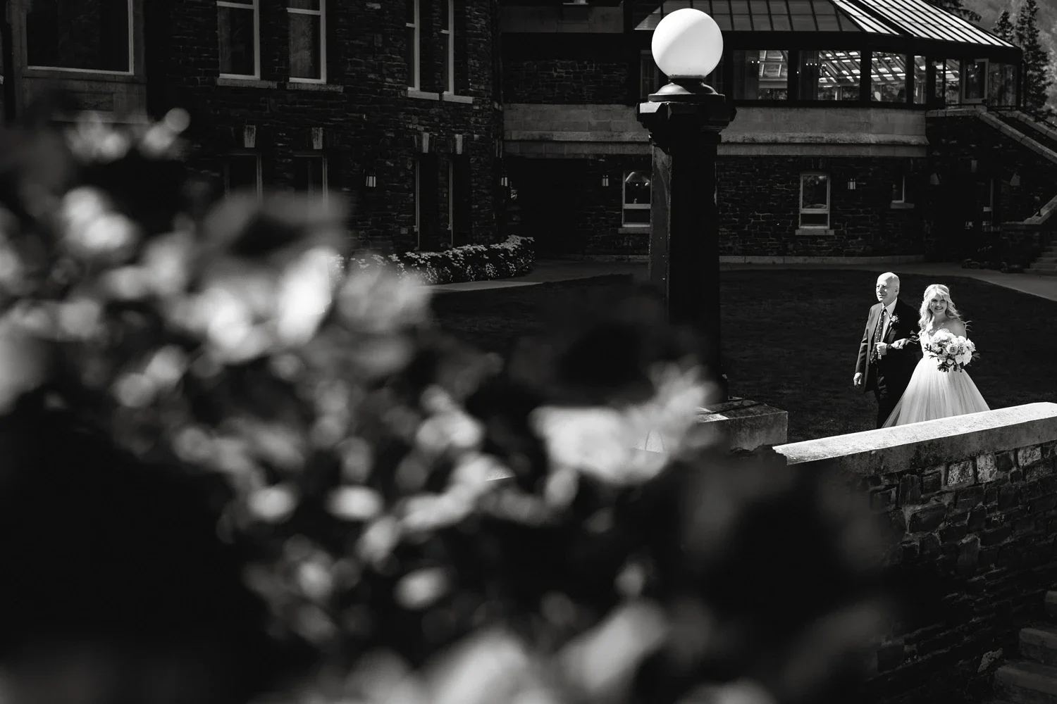 A bride in a white wedding dress holding a bouquet, walking with an older man in a suit outdoors near a brick building with a large lamp post in the foreground.