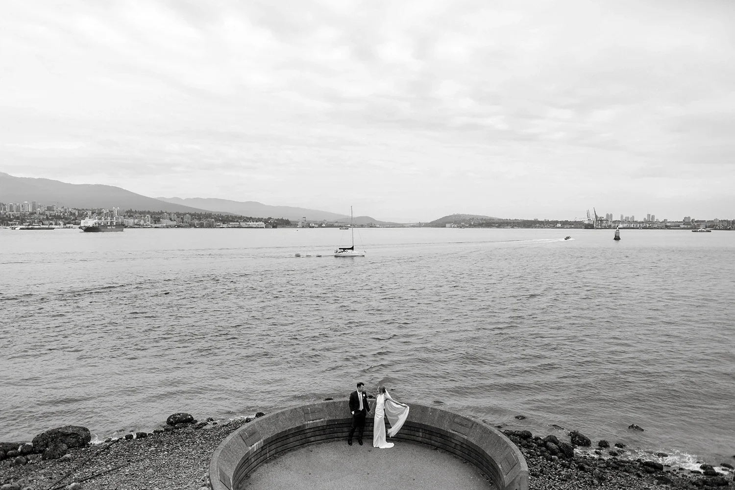 Black and white photo of a couple in wedding attire standing on a rounded viewing platform by the water, with boats and a city skyline in the background.