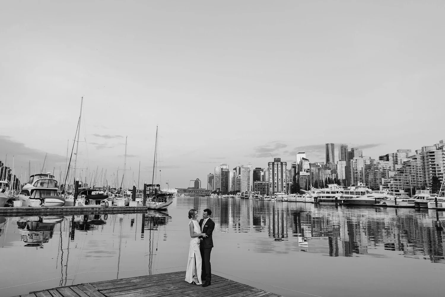 A couple dressed in formal attire standing on a wooden dock by the water in a city marina with boats and yachts, with a skyline of modern high-rise buildings in the background, in black and white.