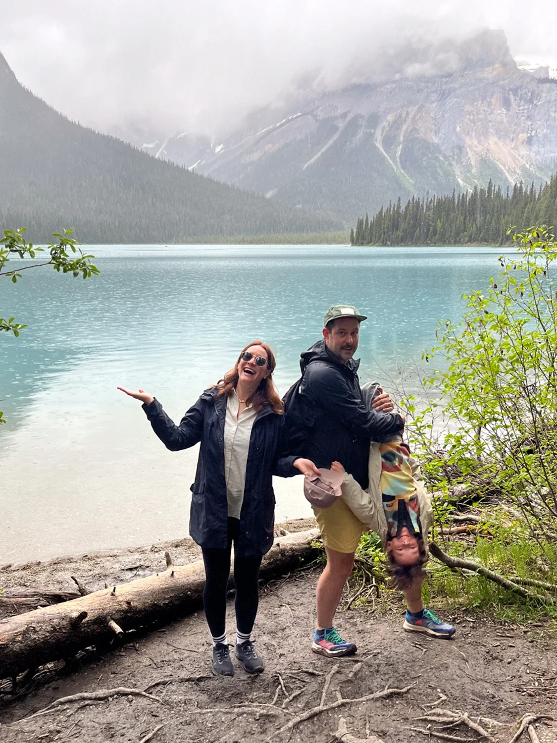 Two adults and a child pose playfully near a lake with mountains and pine trees in the background. One adult stands smiling with arms outstretched, and the other adult is holding the child upside down. The scene is outdoors with overcast weather.