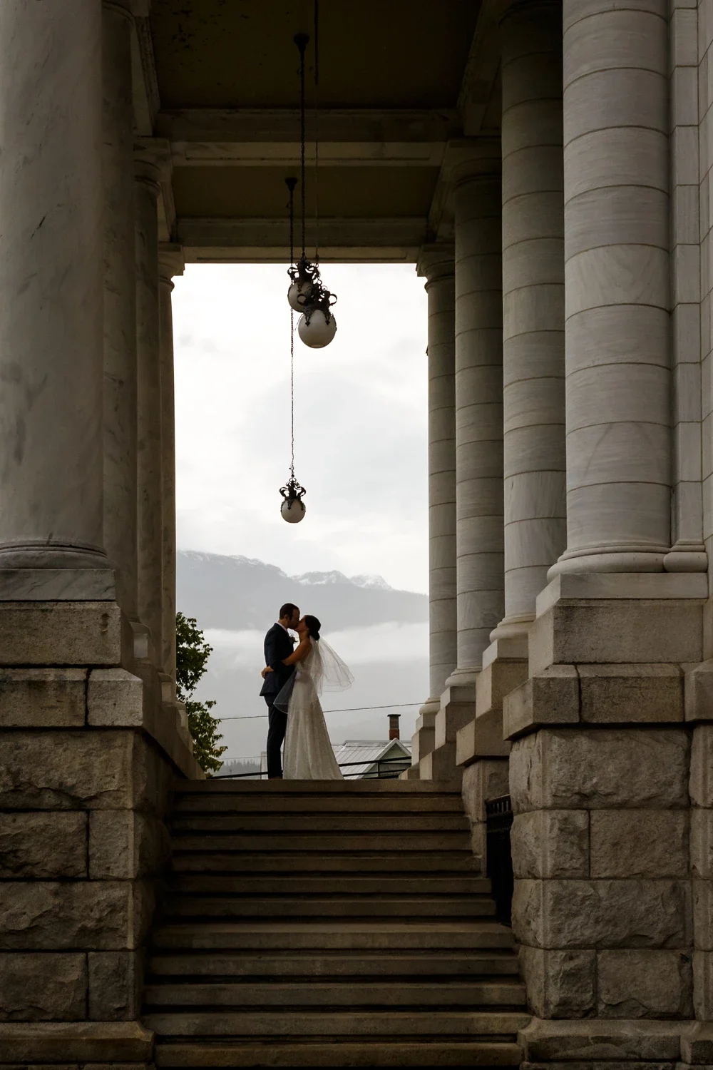 A bride and groom standing on steps in front of a classical building with large columns, sharing a kiss under a cloudy sky.