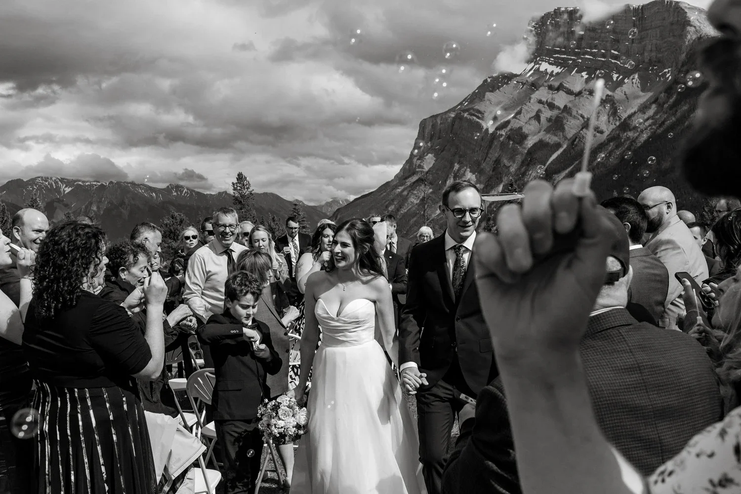Black and white photo of a wedding ceremony outdoors with mountains and cloudy sky in the background. The bride and groom are walking together, holding hands, surrounded by guests. A person in the foreground is blowing bubbles.